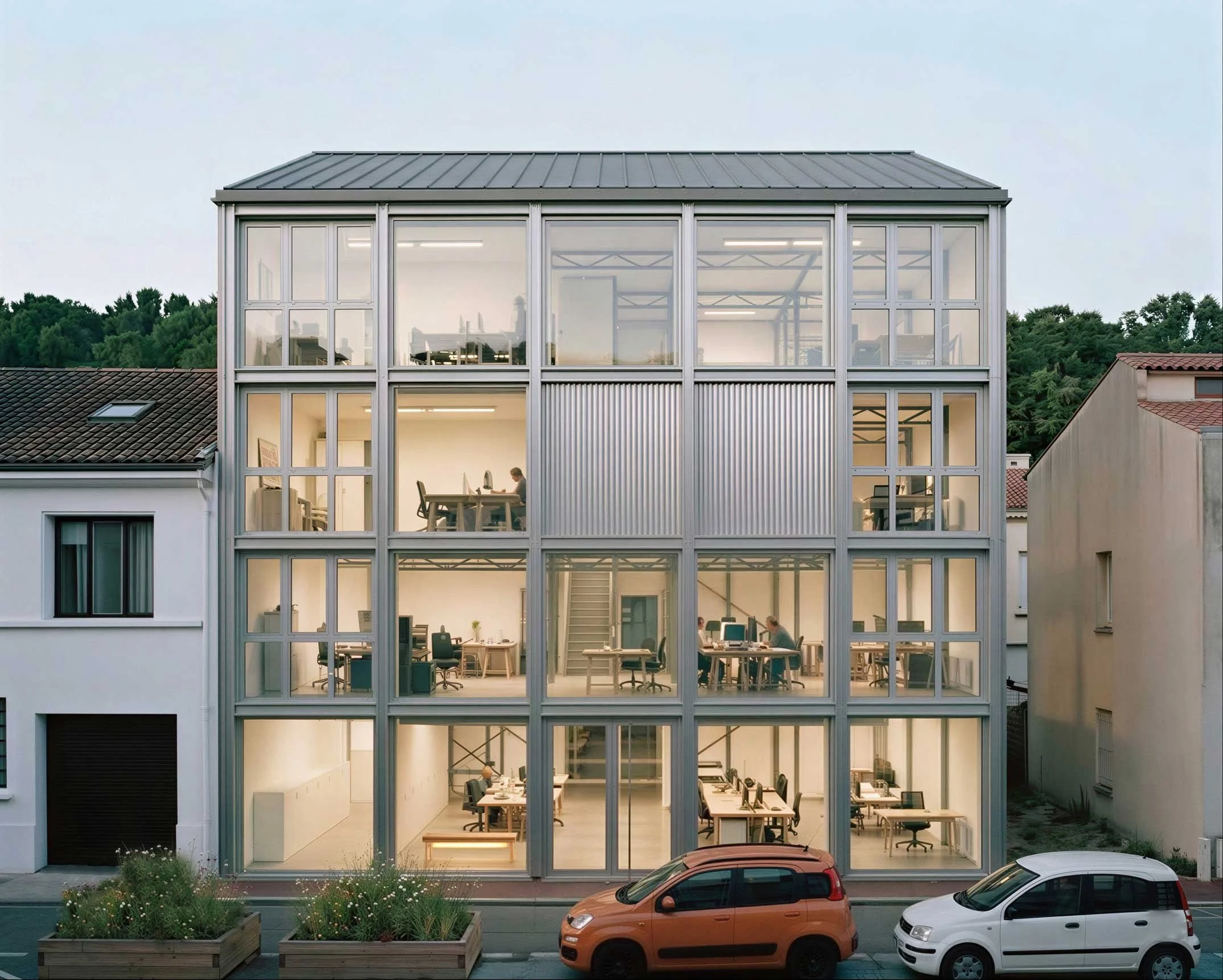 Bâtiment moderne avec grandes fenêtres vitrées laissant voir des employés travaillant à l'intérieur, voitures garées devant, végétation en premier plan, ciel clair en arrière-plan.