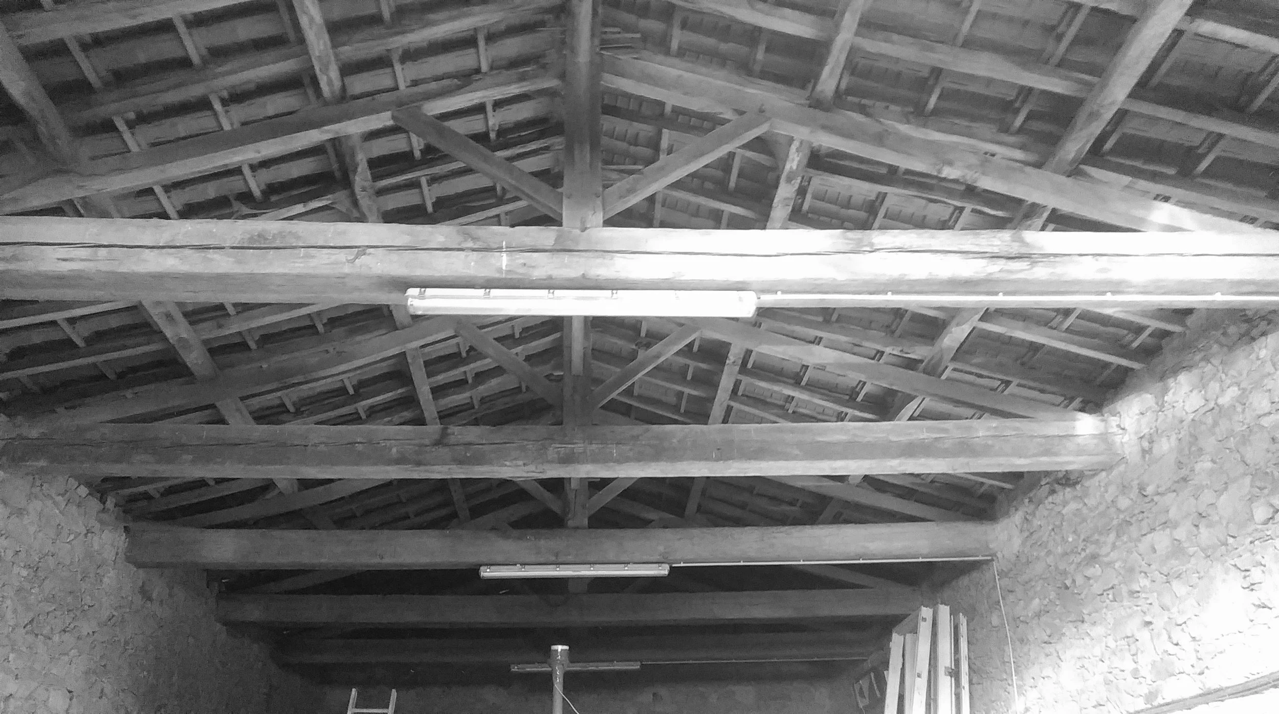 Toit en bois en construction, avec des poutres et une structure de support, toiture en tuiles, murs en pierre. Underground concré et matériel de construction visible.