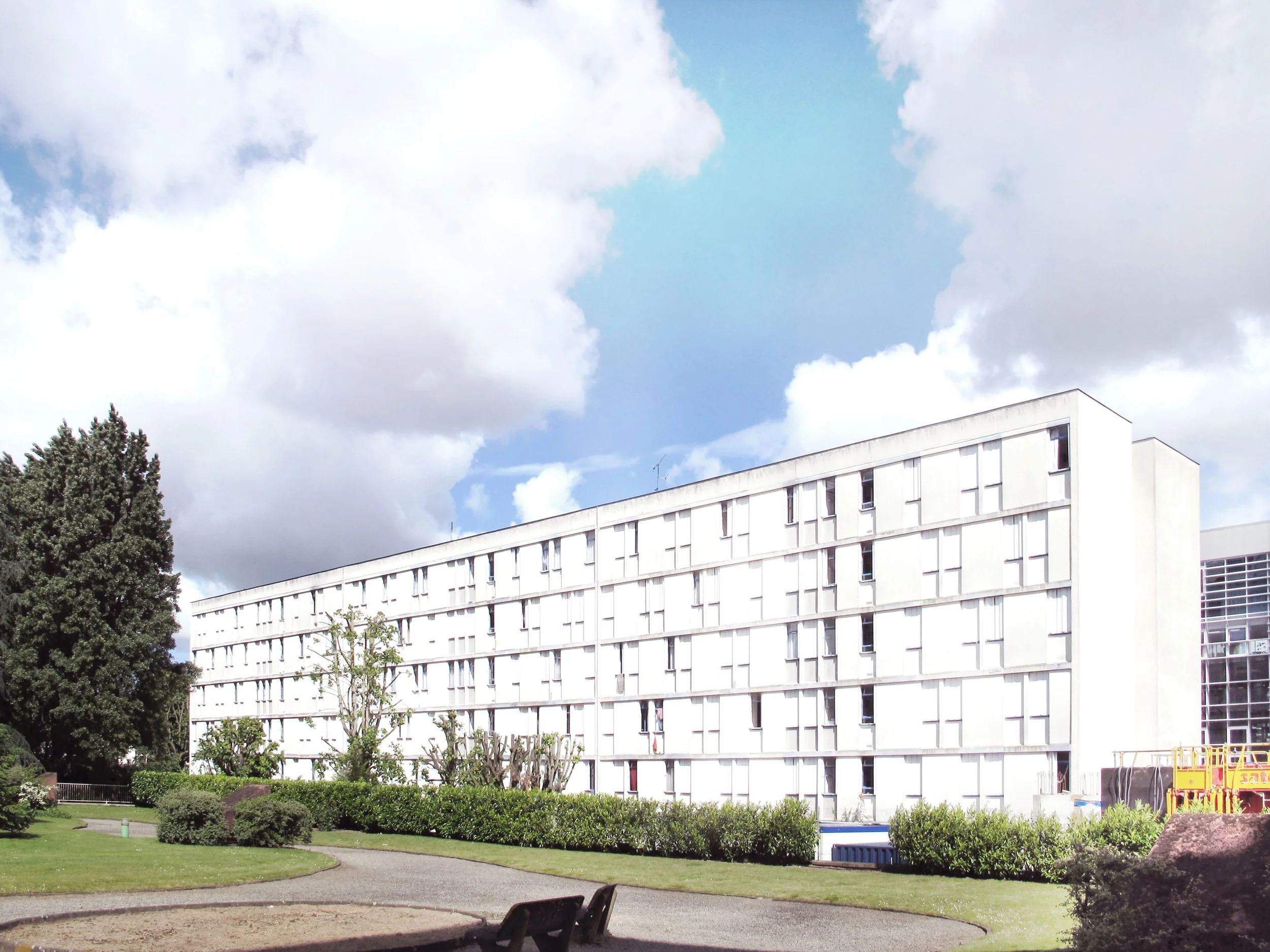 Un bâtiment blanc moderne avec plusieurs fenêtres, un ciel bleu avec des nuages, un arbre feuillu et un espace vert avec un sentier et deux bancs dans un parc.