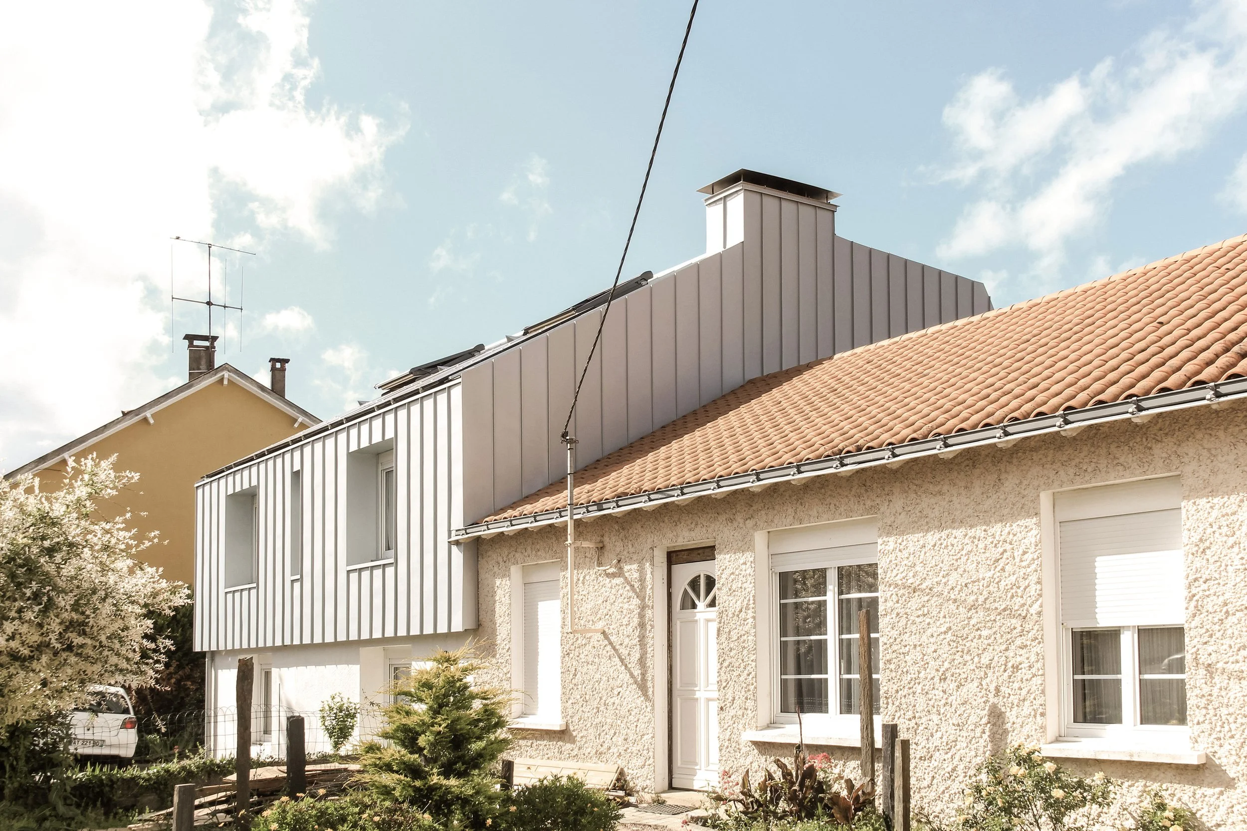 Façade d'une maison avec un toit en tuiles, fenêtres blanches, et un balcon en bois. Il y a un arbre et un véhicule blanc dans le jardin, sous un ciel partiellement nuageux.