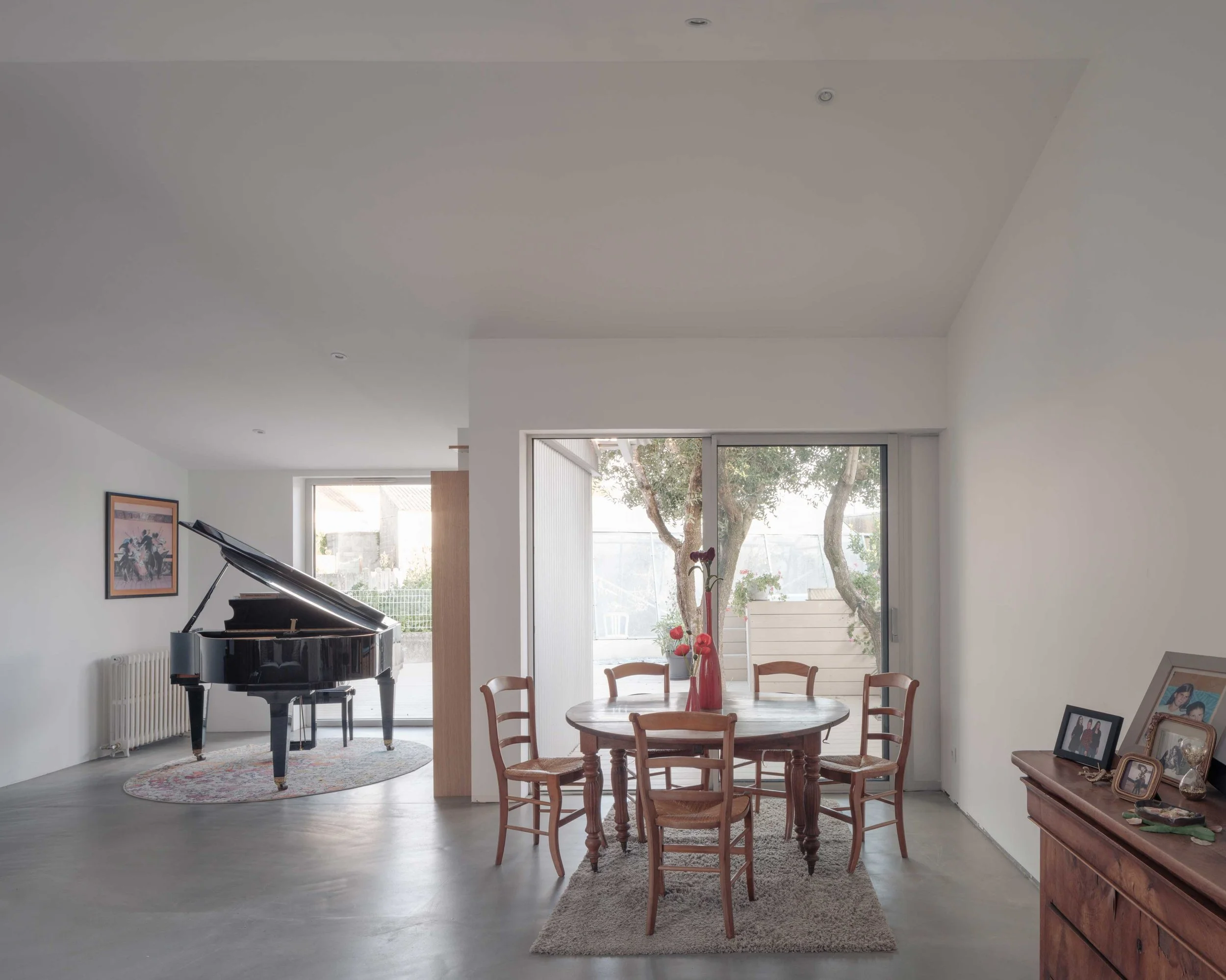 Intérieur d'une pièce avec une grande fenêtre laissant entrer la lumière naturelle, un piano à queue noir dans un coin, une table ronde en bois entourée de chaises en bois, un tapis beige, des cadres photo posés sur un meuble en bois, des plantes en 