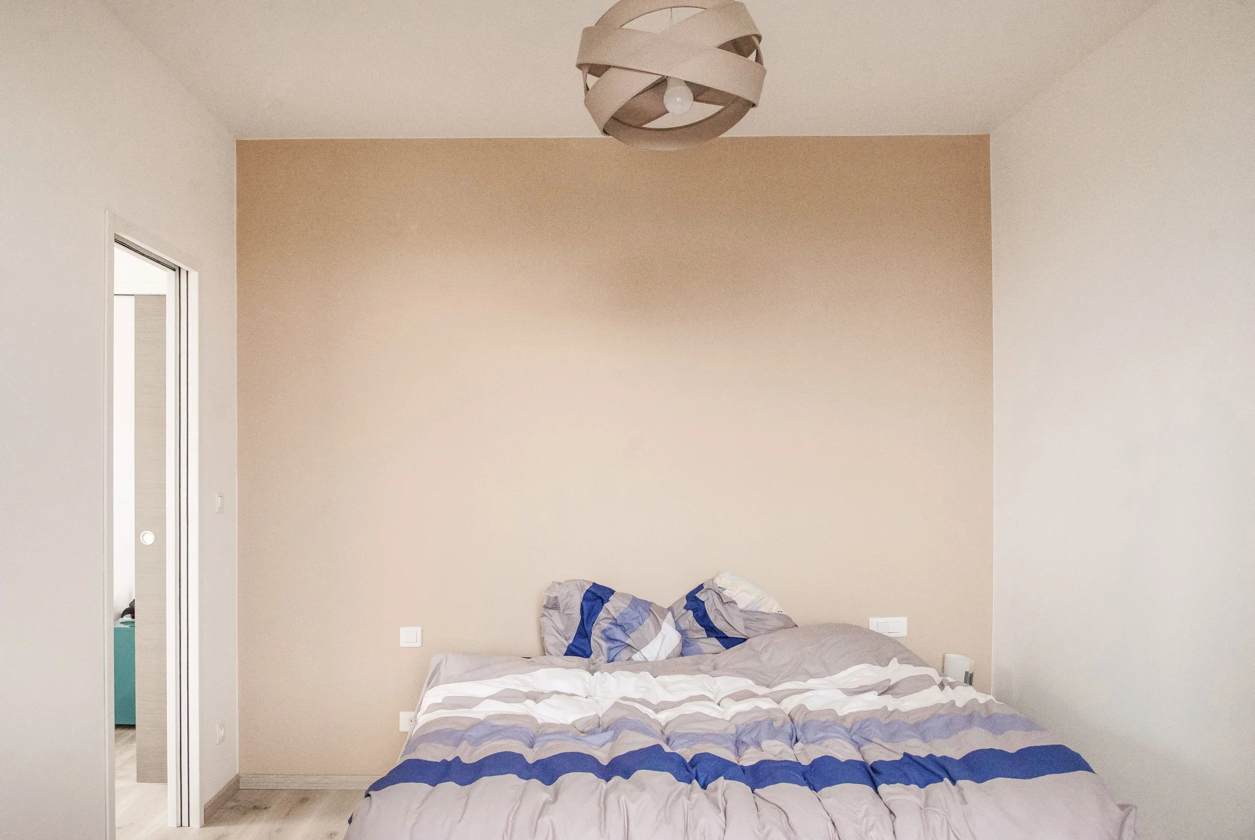 Chambre avec un lit non fait, un mur beige, une fenêtre, et une porte menant à une autre pièce.