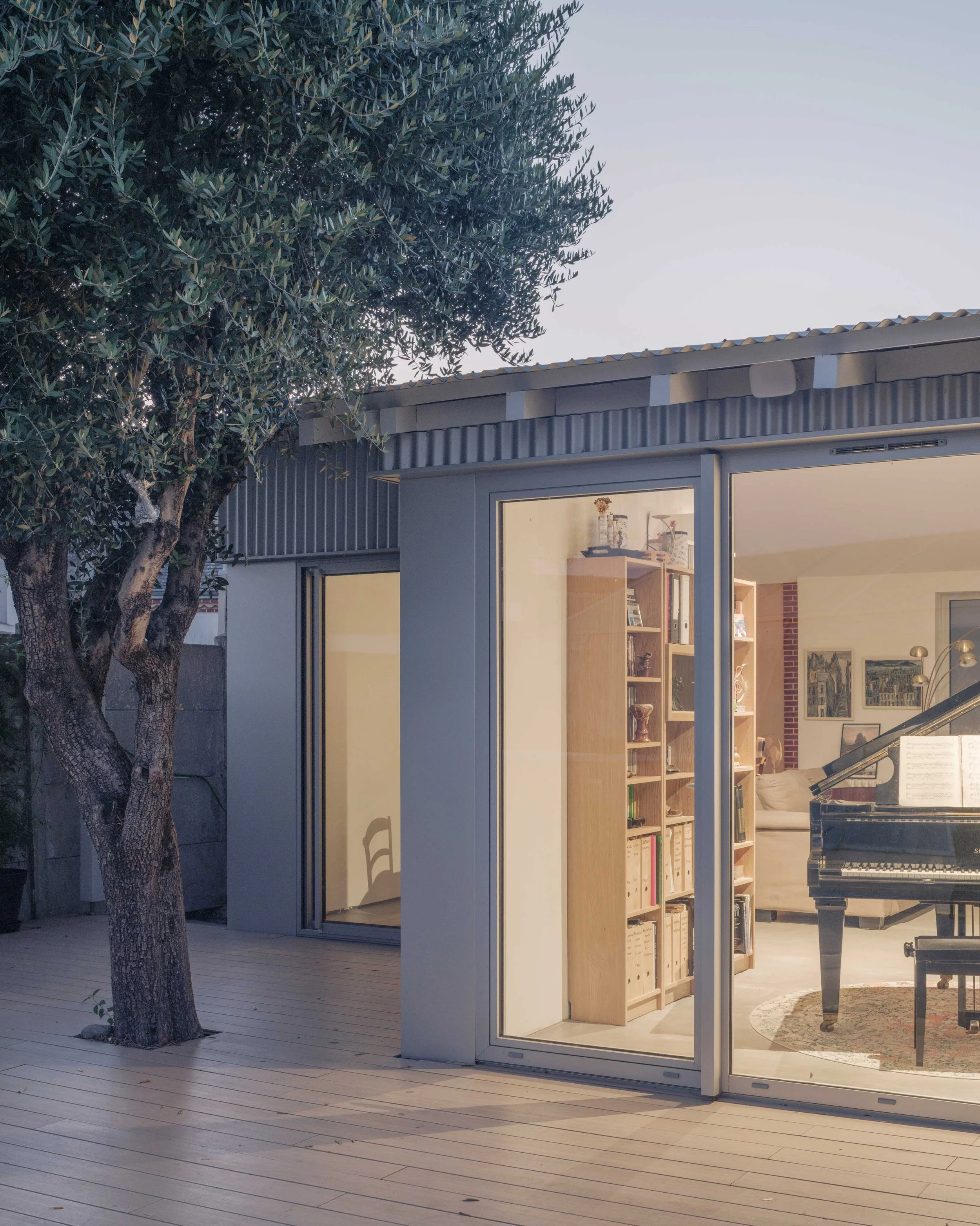 Une maison moderne avec une façade en verre, à côté d'un arbre, et une étagère visible à travers la fenêtre intérieure.