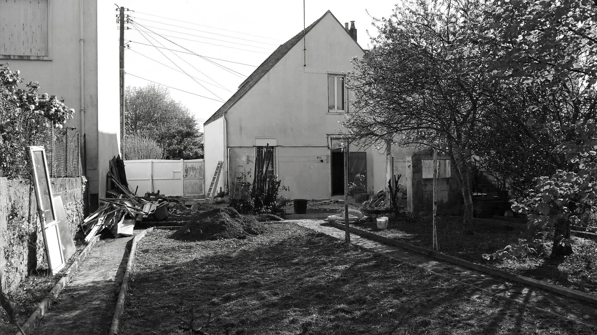 Une maison en construction ou en rénovation dans un jardin, avec des outils, des matériaux, et des arbres environnants, en noir et blanc.