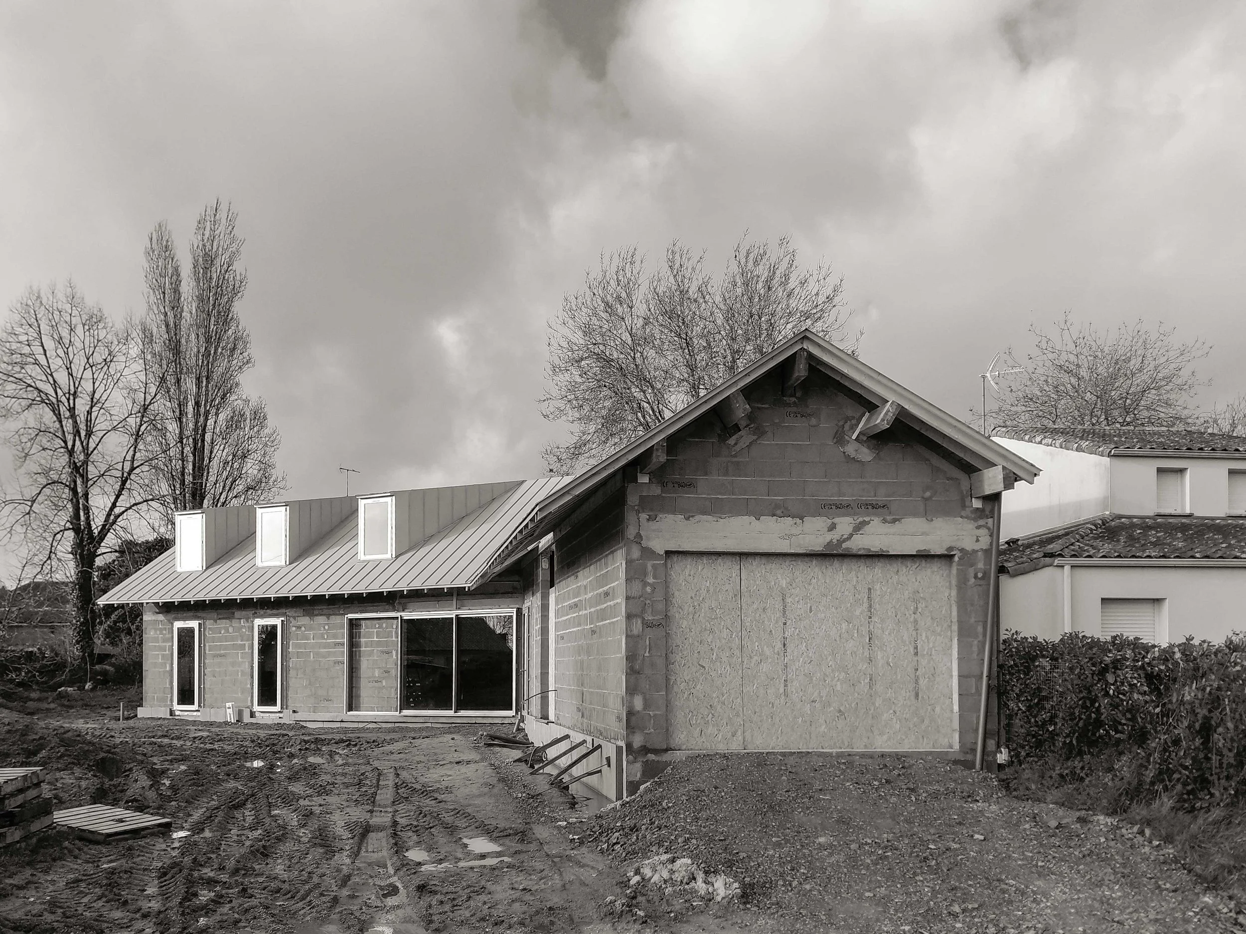 Une maison en construction avec murs en blocs de béton, fenêtres installées, et un toit en métal. Le terrain autour est en cours de terrassement avec des traces de mouvement de terre.