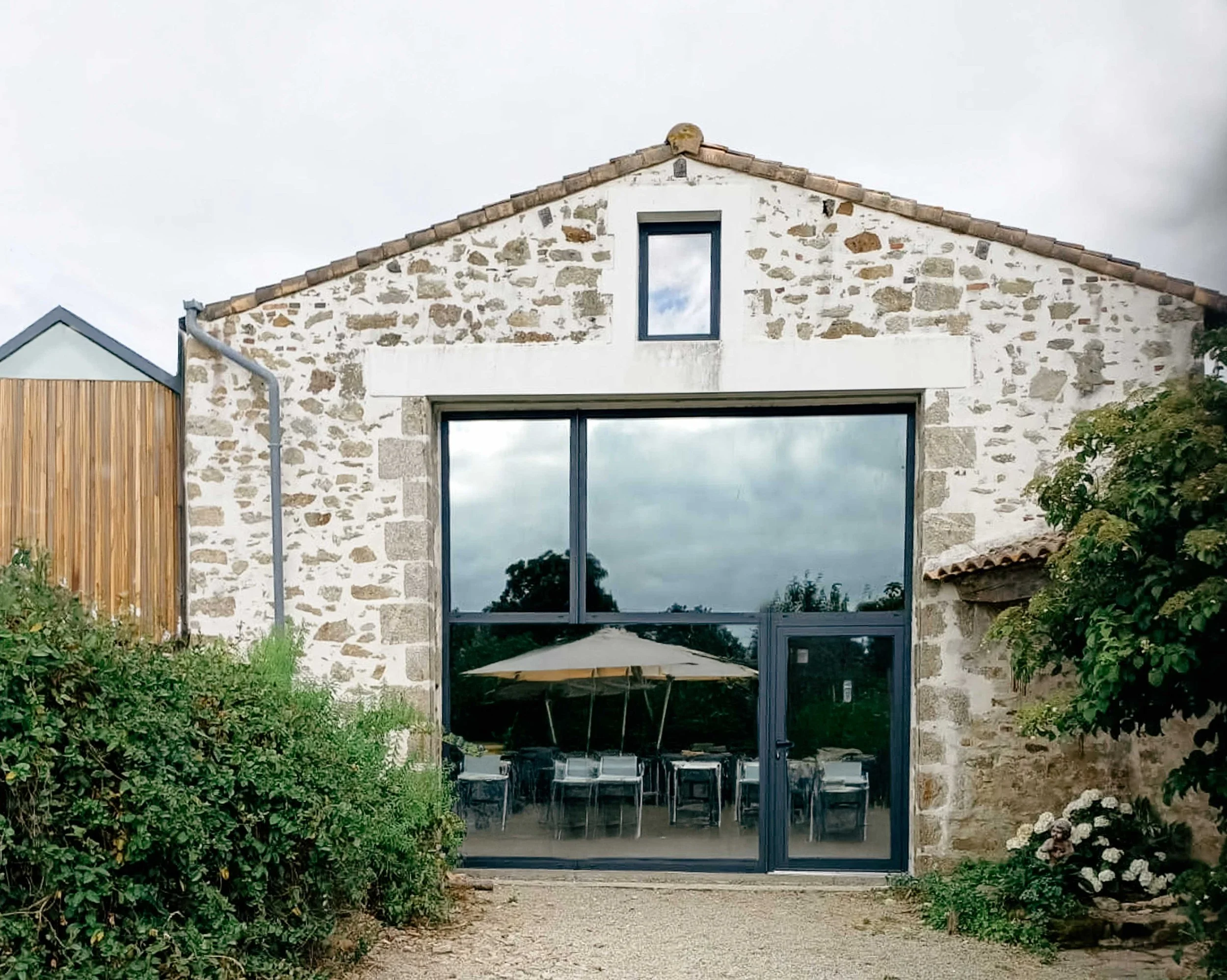 Travée d'une maison en pierre avec grandes fenêtres, une porte vitrée et une toiture en tuiles, entourée de végétation et de galets.