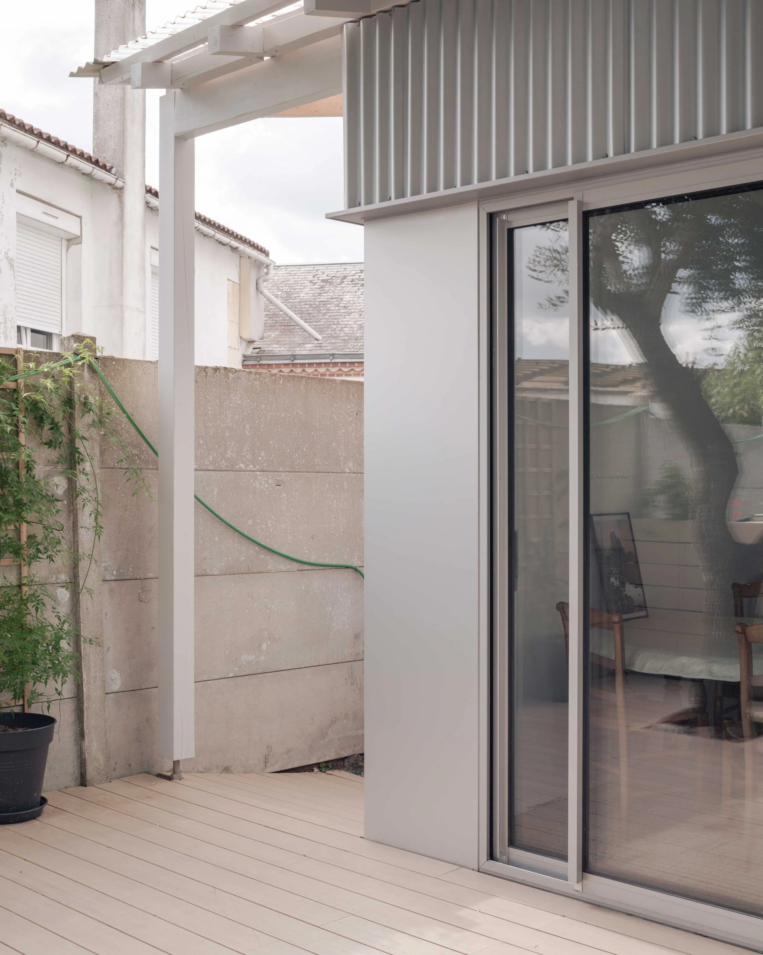 Terrasse en bois avec une porte-fenêtre en verre et une structure en métal, vue d'un espace extérieur d'une maison avec mur en béton et plantes en pot.