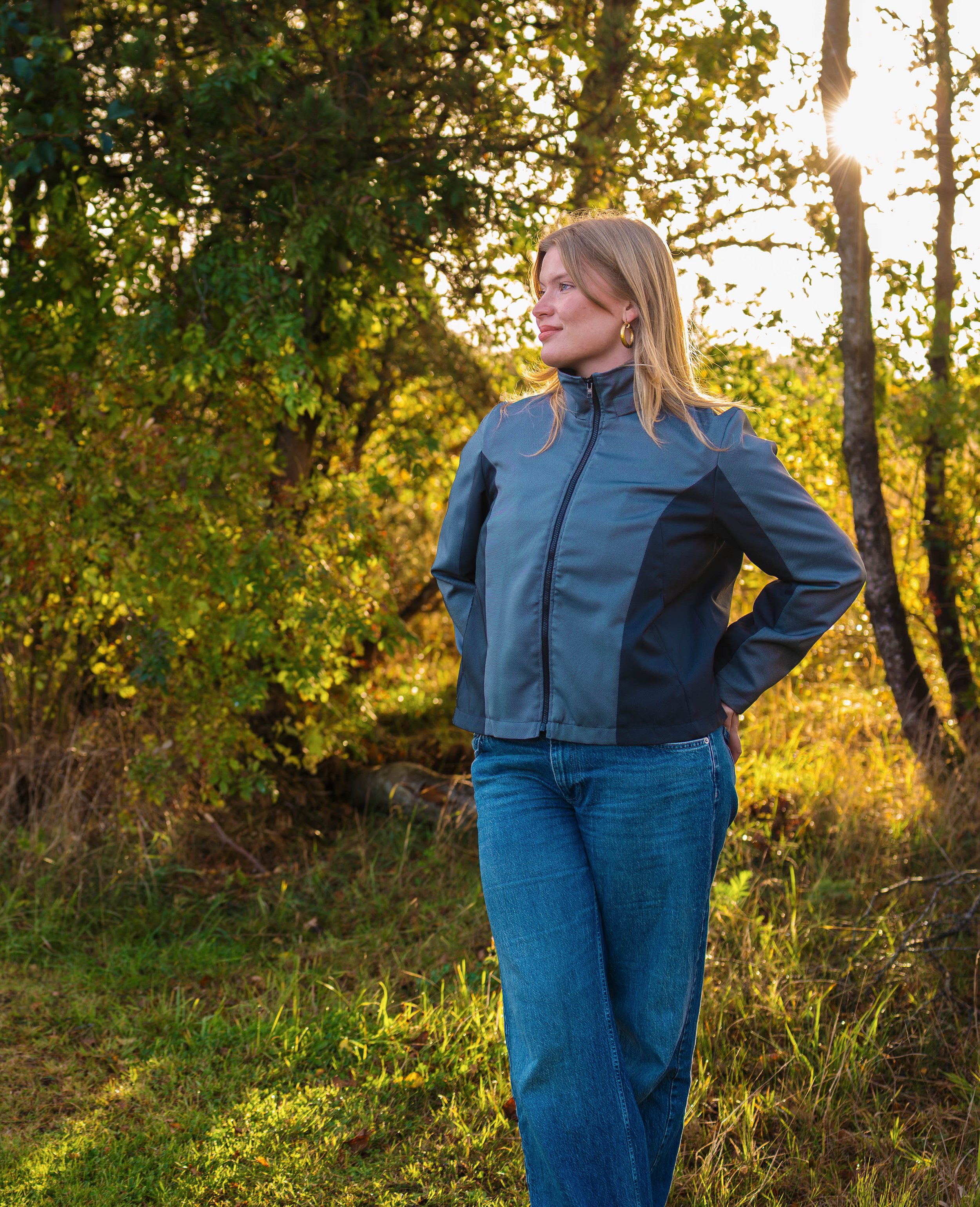 A woman with blonde hair stands outdoors in a forested area during sunset, wearing a Vrang Vision jacket and blue jeans.