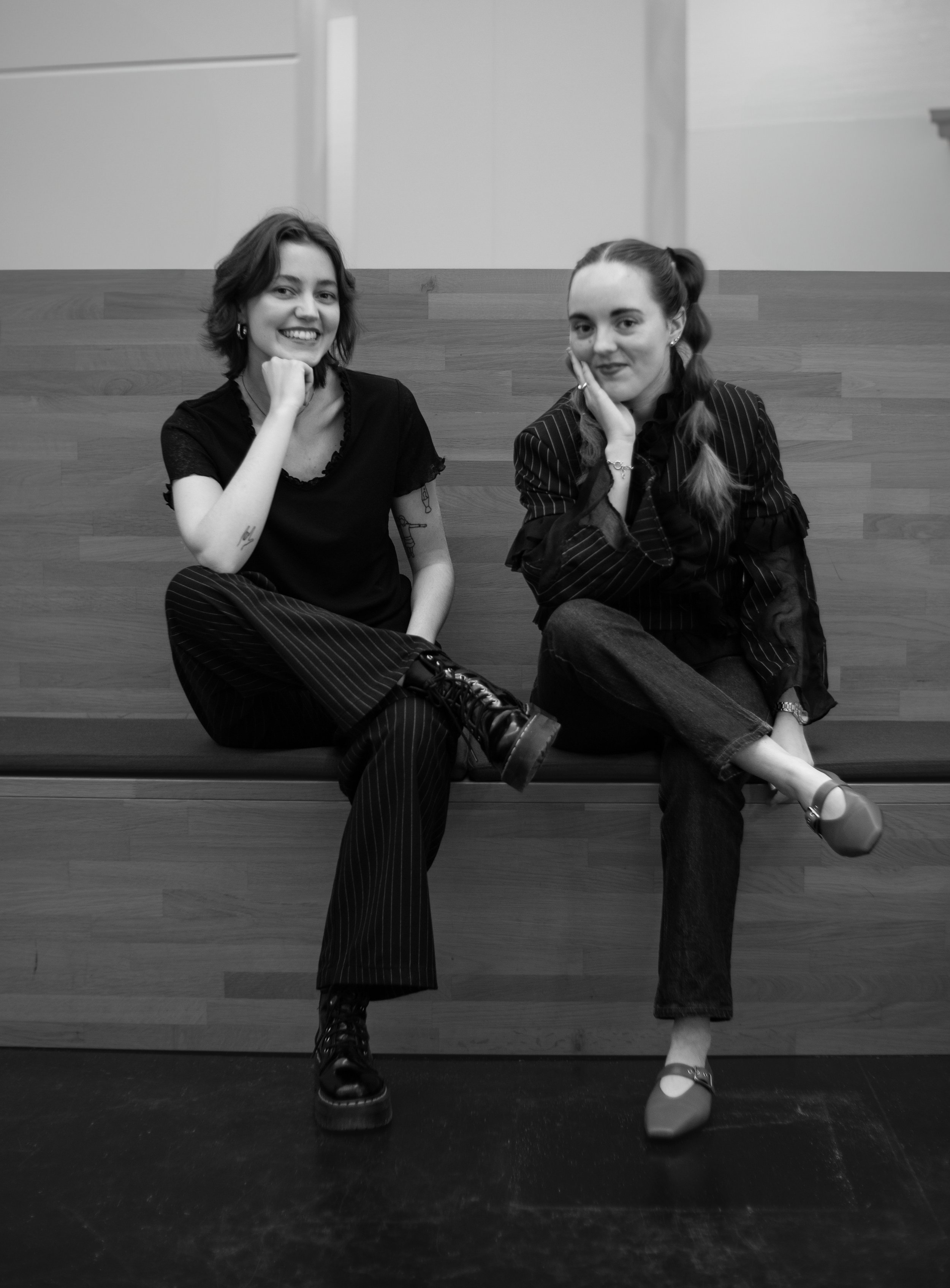 The Vrang Vision founder team: Two women sitting on a wooden bench, smiling and posing for the camera in a modern interior setting.