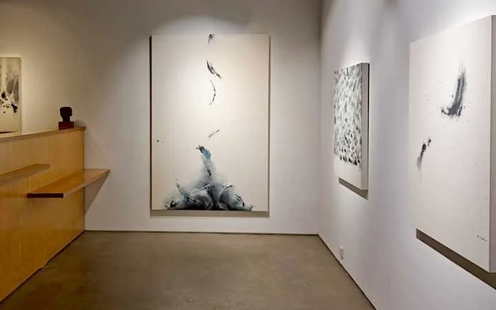An art gallery with three abstract paintings on white walls. The paintings feature minimalistic, fluid brushstrokes and patterns.