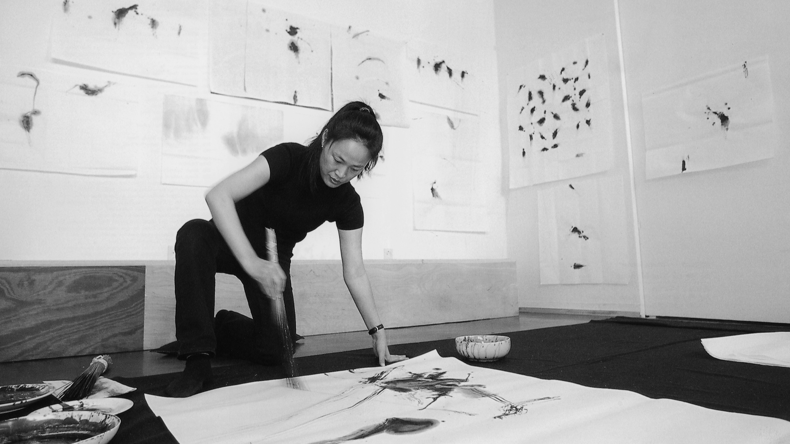 Woman creating traditional Asian ink painting on large sheet of paper, surrounded by brushes and ink bowls, with abstract artwork on the wall behind her.
