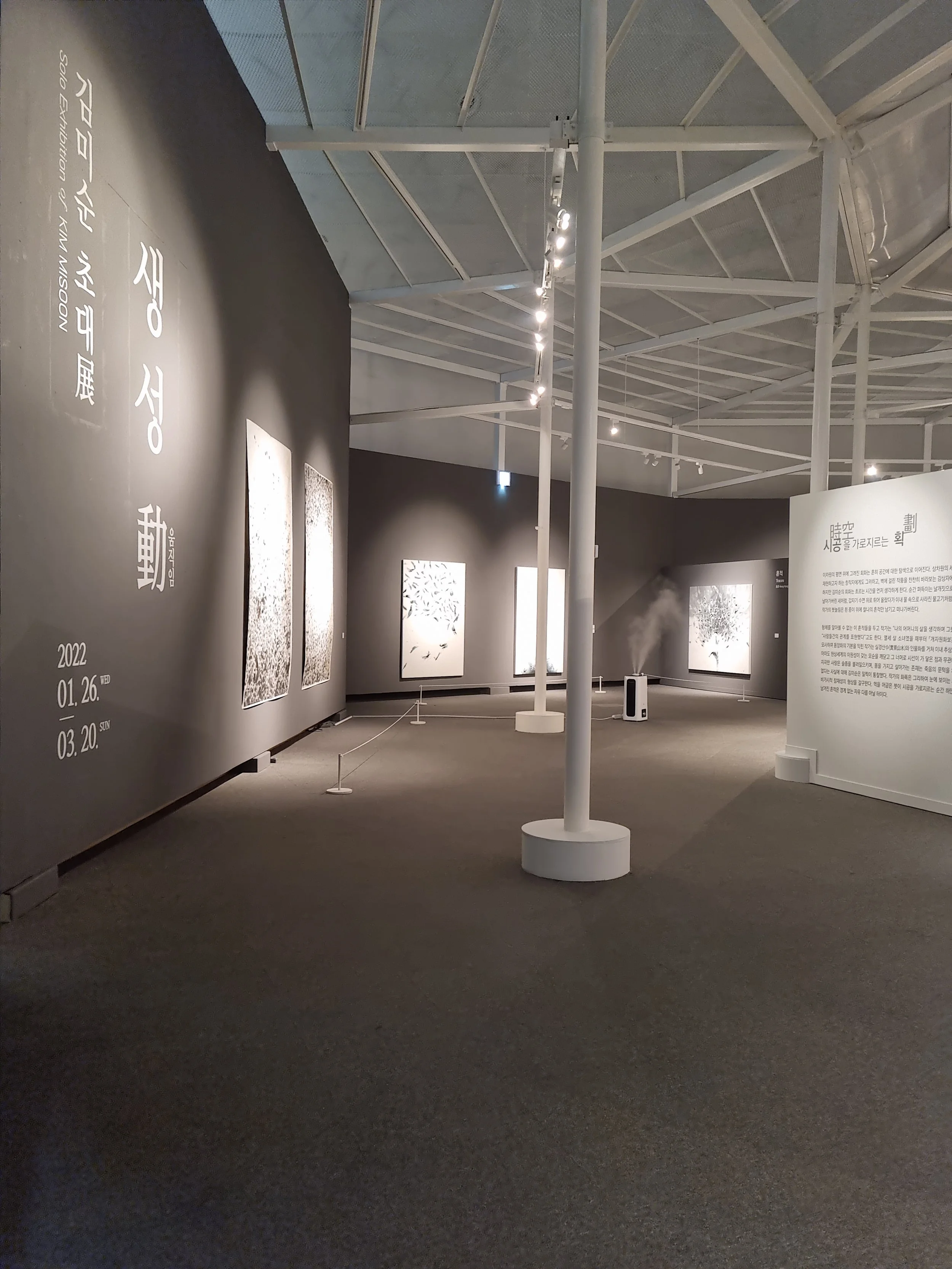 Interior of an art exhibition with several black and white paintings displayed on dark walls and a large white informational panel.