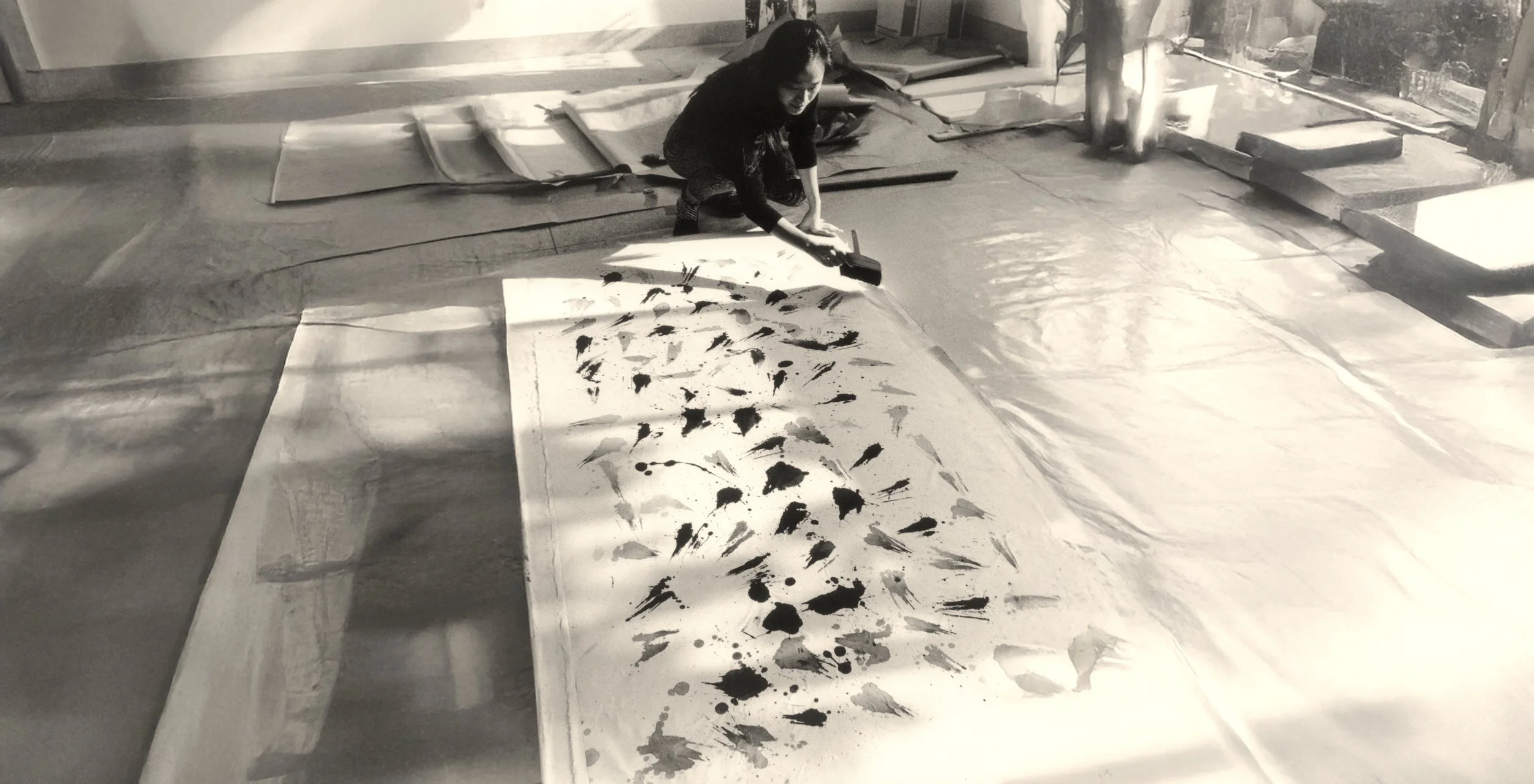 A woman kneeling on the floor, painting a large canvas with abstract black ink splashes.