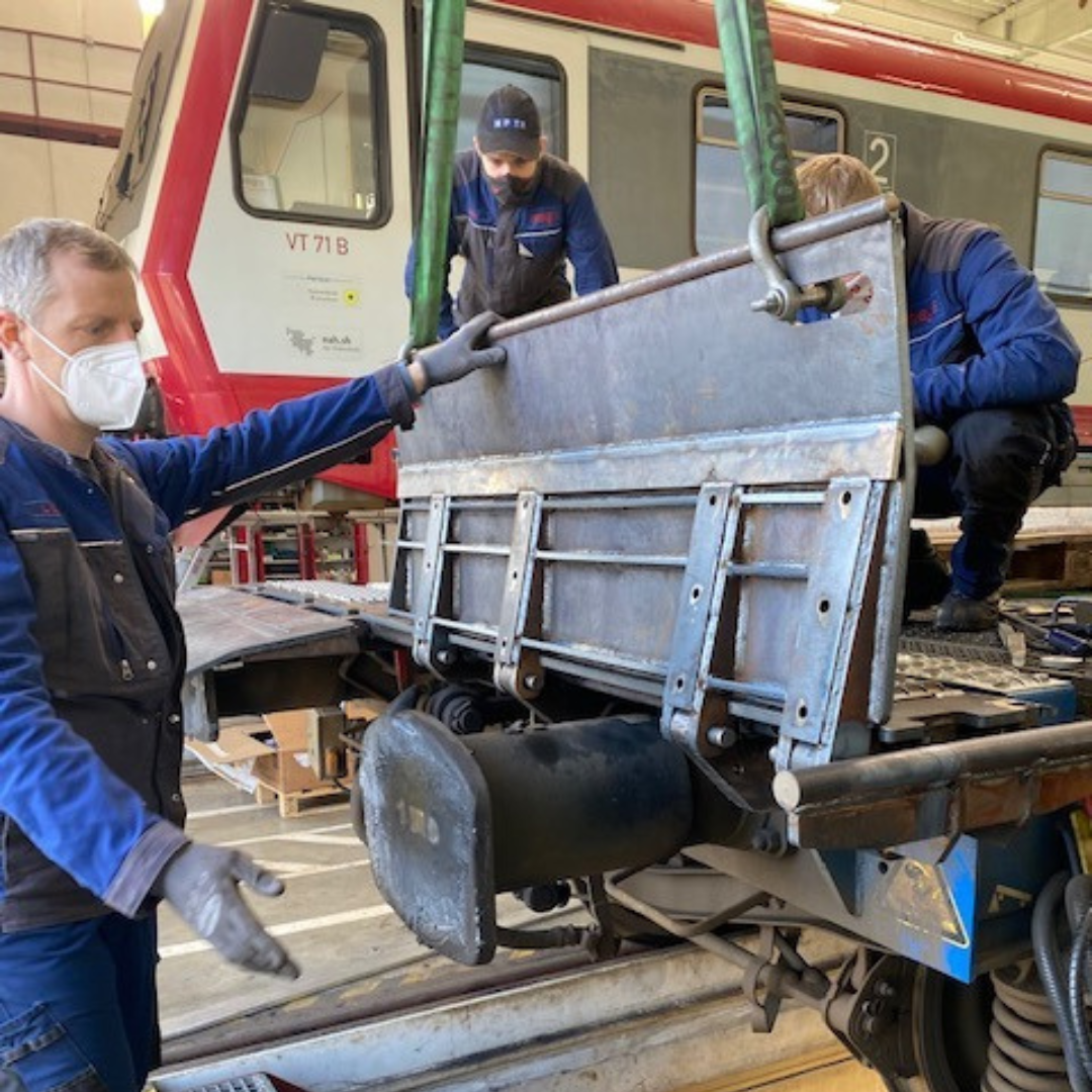 Drei Arbeiter in Schutzkleidung und Masken arbeiten an einer Eisenbahn, einer steht seitlich neben den Schienen, die anderen zwei sind am vorderen Teil der Eisenbahn beschäftigt.