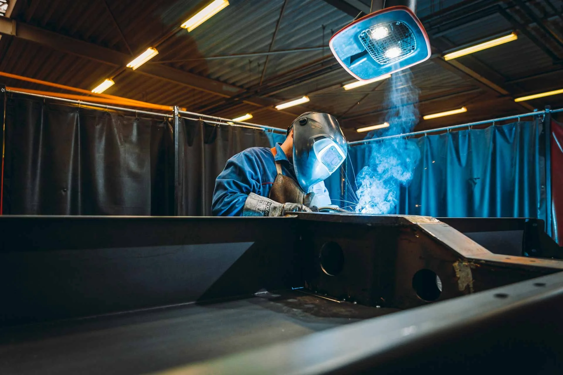 Schweißer arbeitet in einer Werkstatt mit Schutzhelm, während Funken fliegen und Rauch aufsteigen.