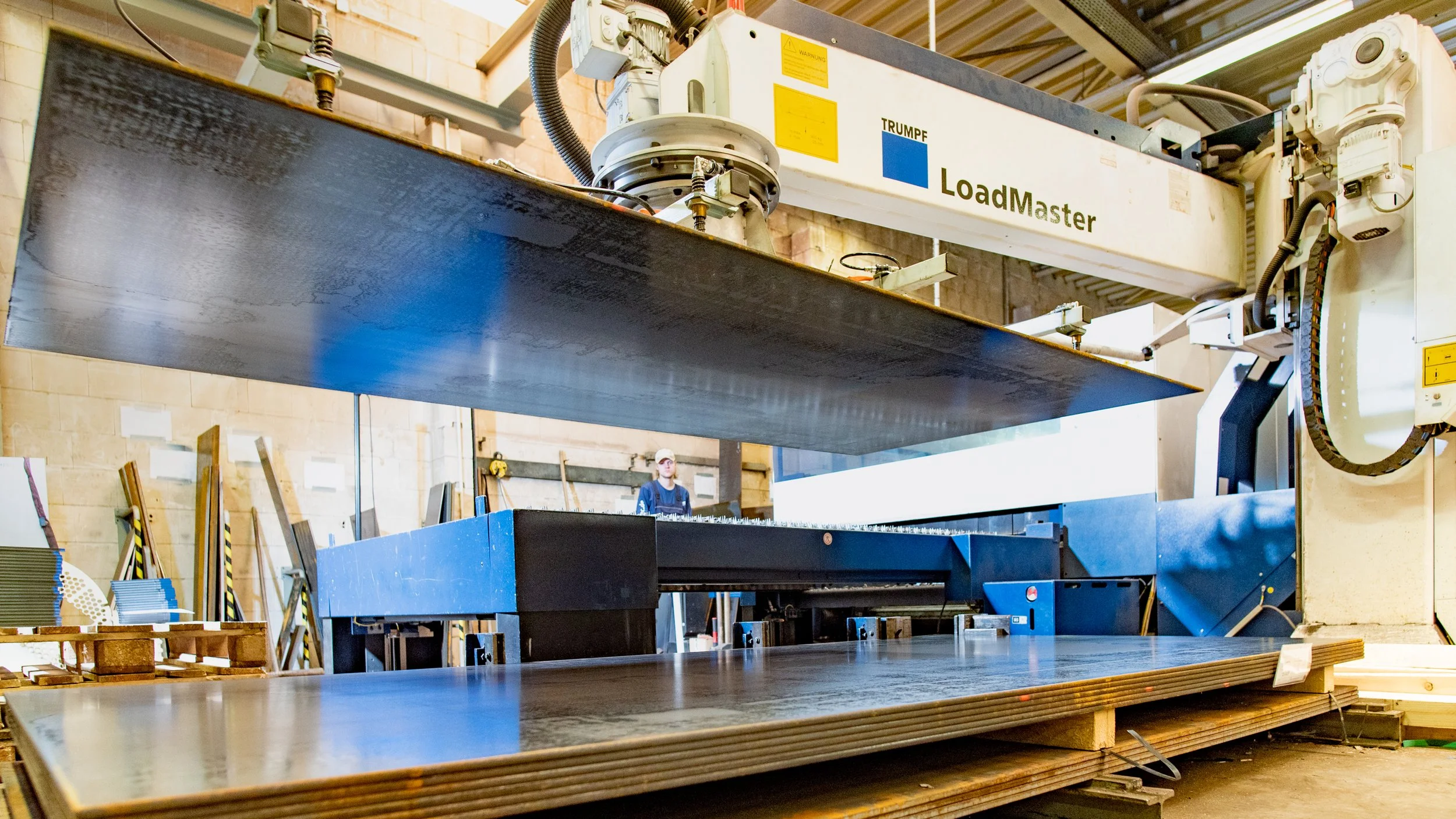 Große industrielle Maschine zum Schneiden oder Bearbeiten von Holzplatten in einer Werkhalle, mit einem Arbeiter im Hintergrund.
