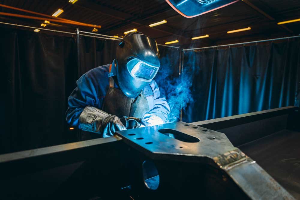 Schweißer bei der Arbeit in einer Werkstatt, umgeben von schwarzen Vorhängen, mit Schutzhelm und abgedeckten Händen.