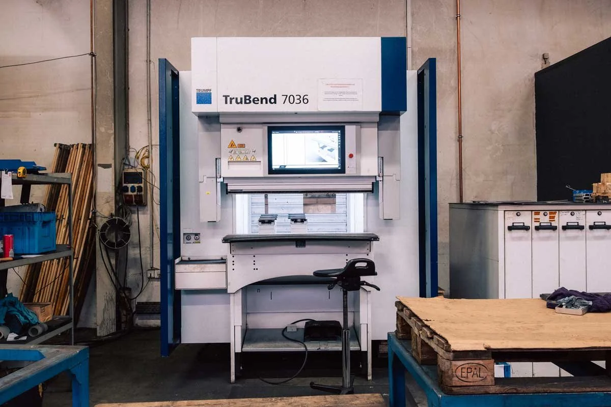Industriewerkstatt mit einer TruBend 7036 Blechbiegemaschine, einem Arbeitstisch, Werkzeugregalen und Kartons.