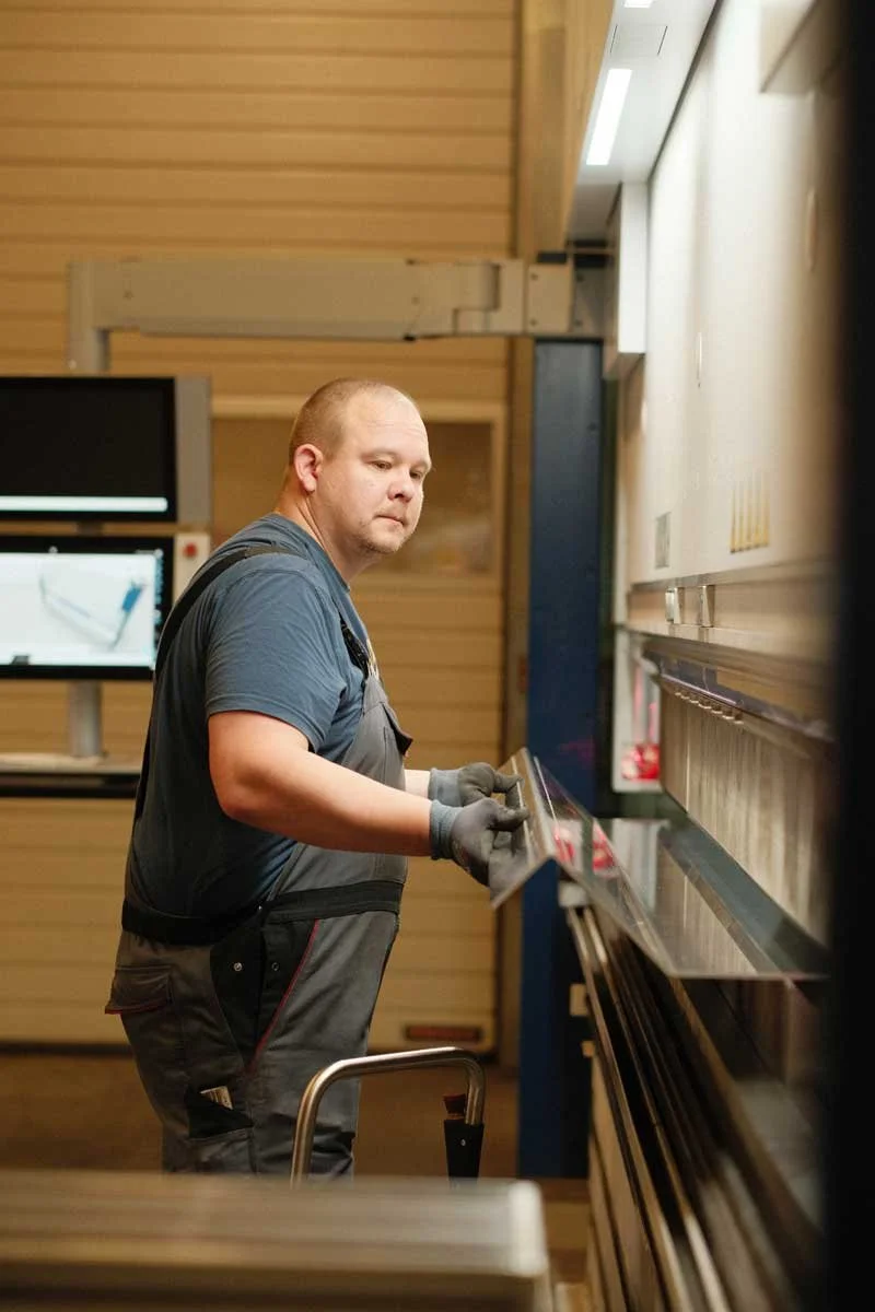 Mann arbeitet in einer Werkstatt an einer Maschine, trägt Handschuhe und eine Schürze.