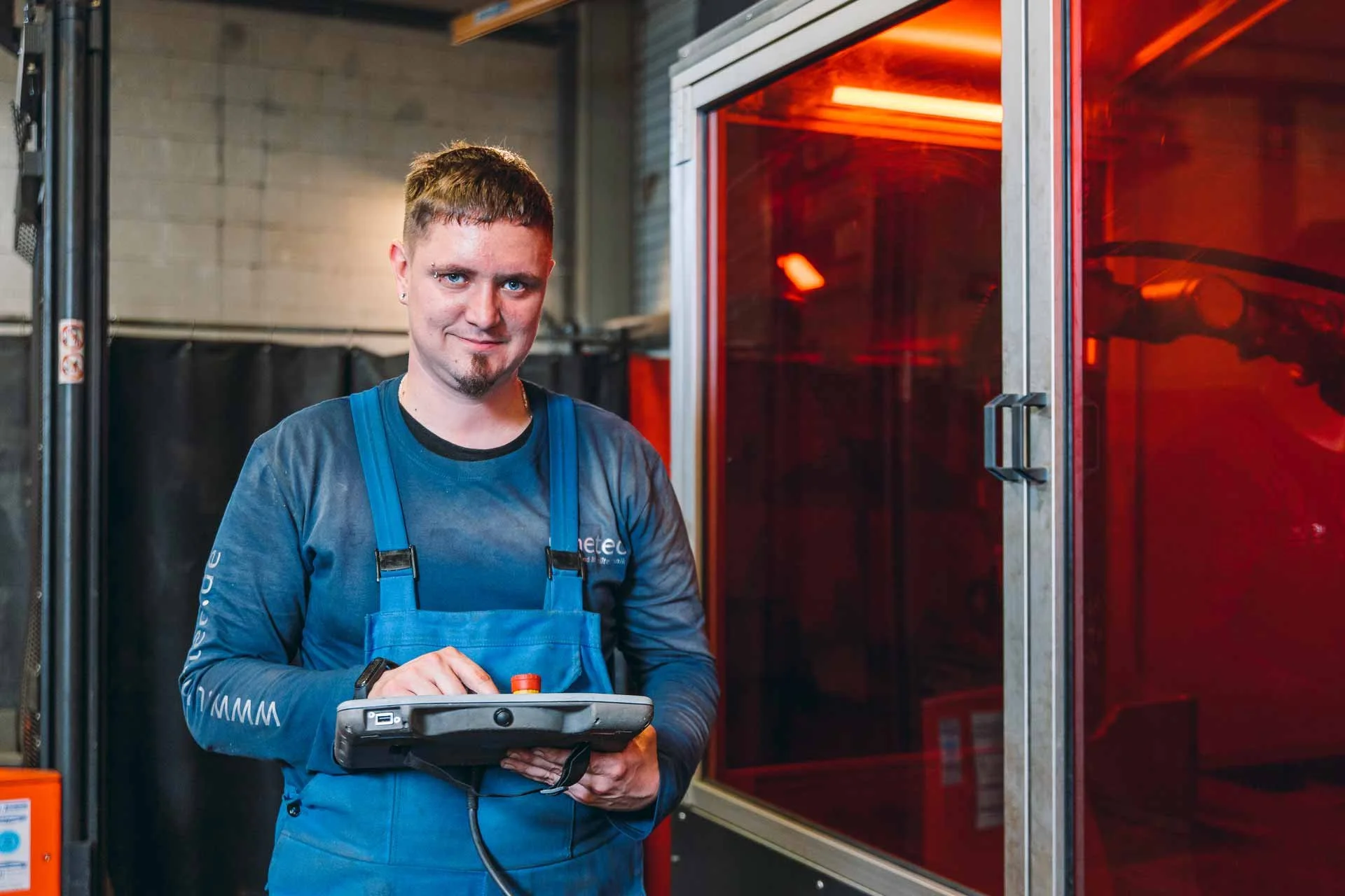 Ein Mann trägt blaue Arbeitskleidung und steht vor einer Maschine mit rotem Licht, hält ein Messgerät in den Händen und sieht in die Kamera.