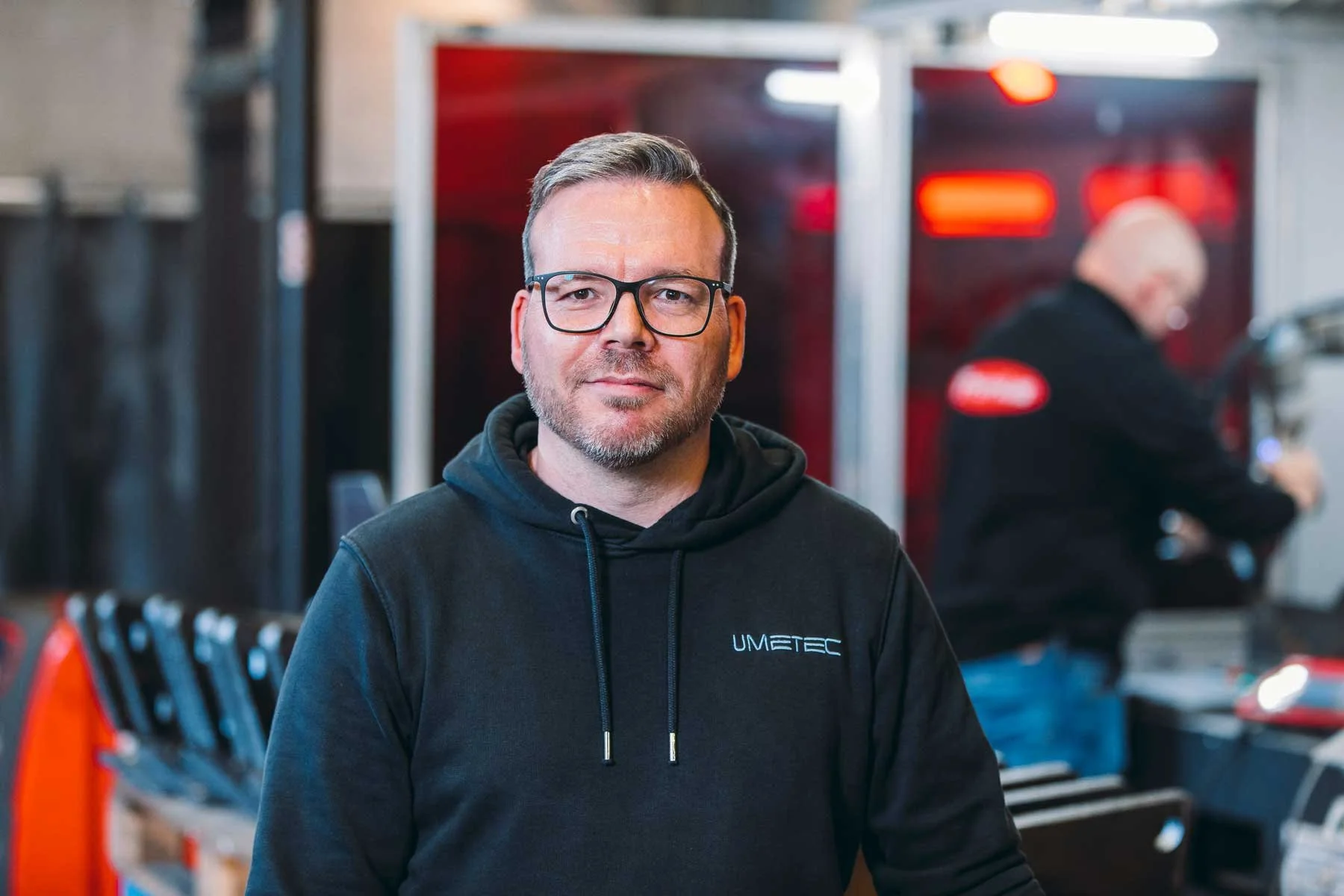 Mann mit Brille in einer Werkstatt mit Werkzeugen und einer Person im Hintergrund.