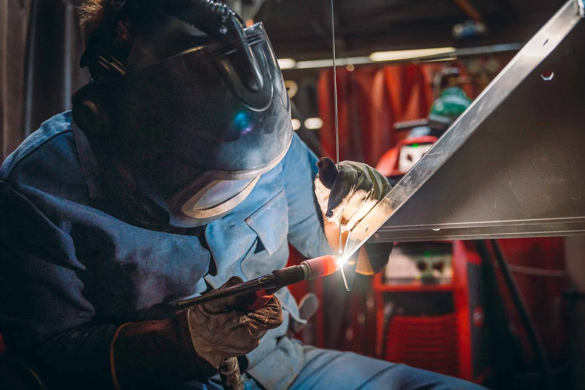 Ein Handwerker schweißt Metall in einer Werkstatt, trägt eine Schutzbrille, einen kohlrabenschwarzen Helm und Arbeitshandschuhe.