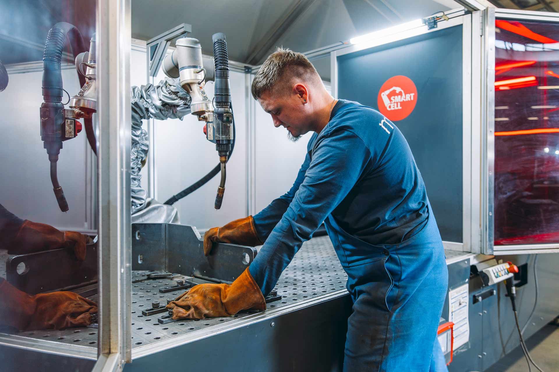 Ein Mann in blauer Arbeitskleidung und braunen Schutzhandschuhen arbeitet mit einer Maschine, die Industrie-Roboterarme und Schneidgeräte enthält, in einer Werkstatt oder Fabrik.
