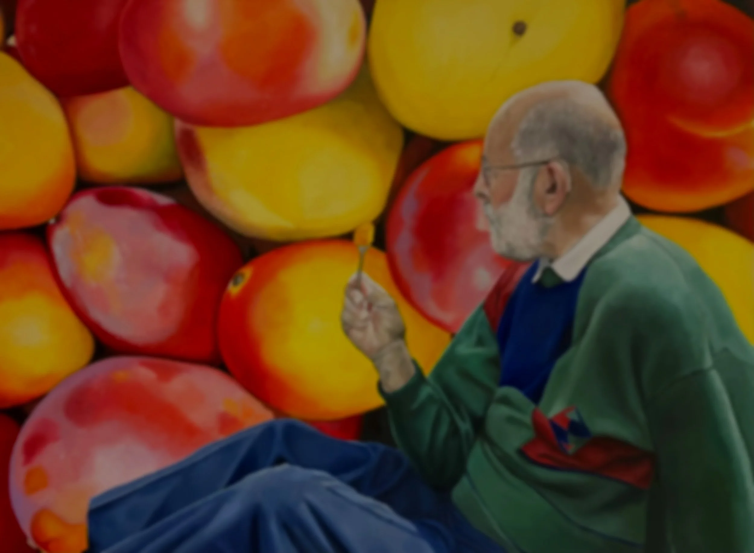 A man with glasses and a beard, wearing a green jacket, sitting on the ground, appears to be painting a mural of apples, which includes large, realistic, red and yellow apples.