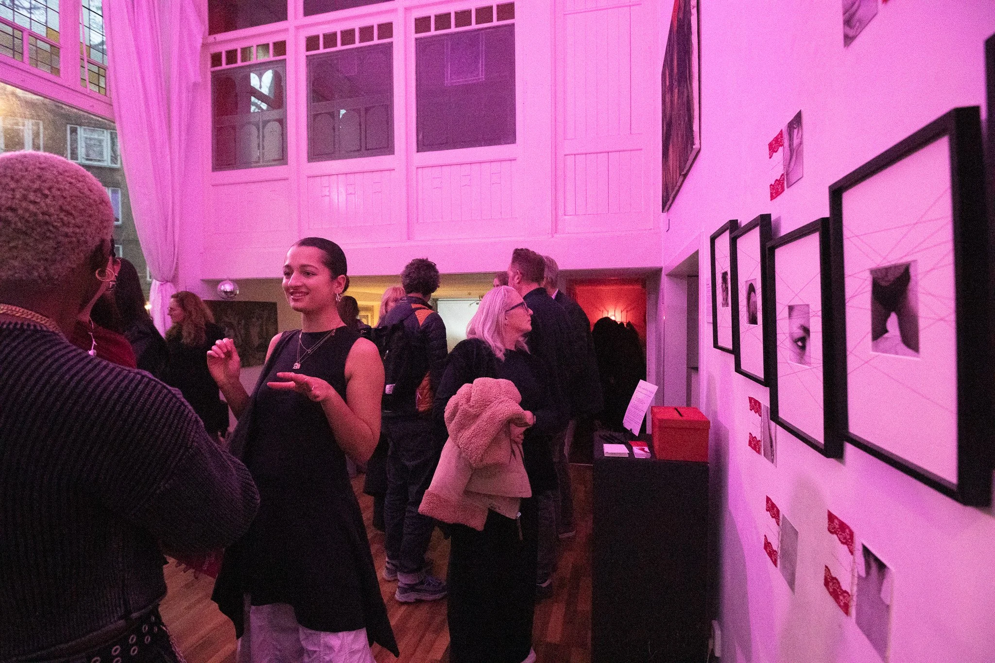 People attending an art gallery exhibit with framed artwork on white walls, pink lighting, and a diverse crowd of visitors engaged in conversation.