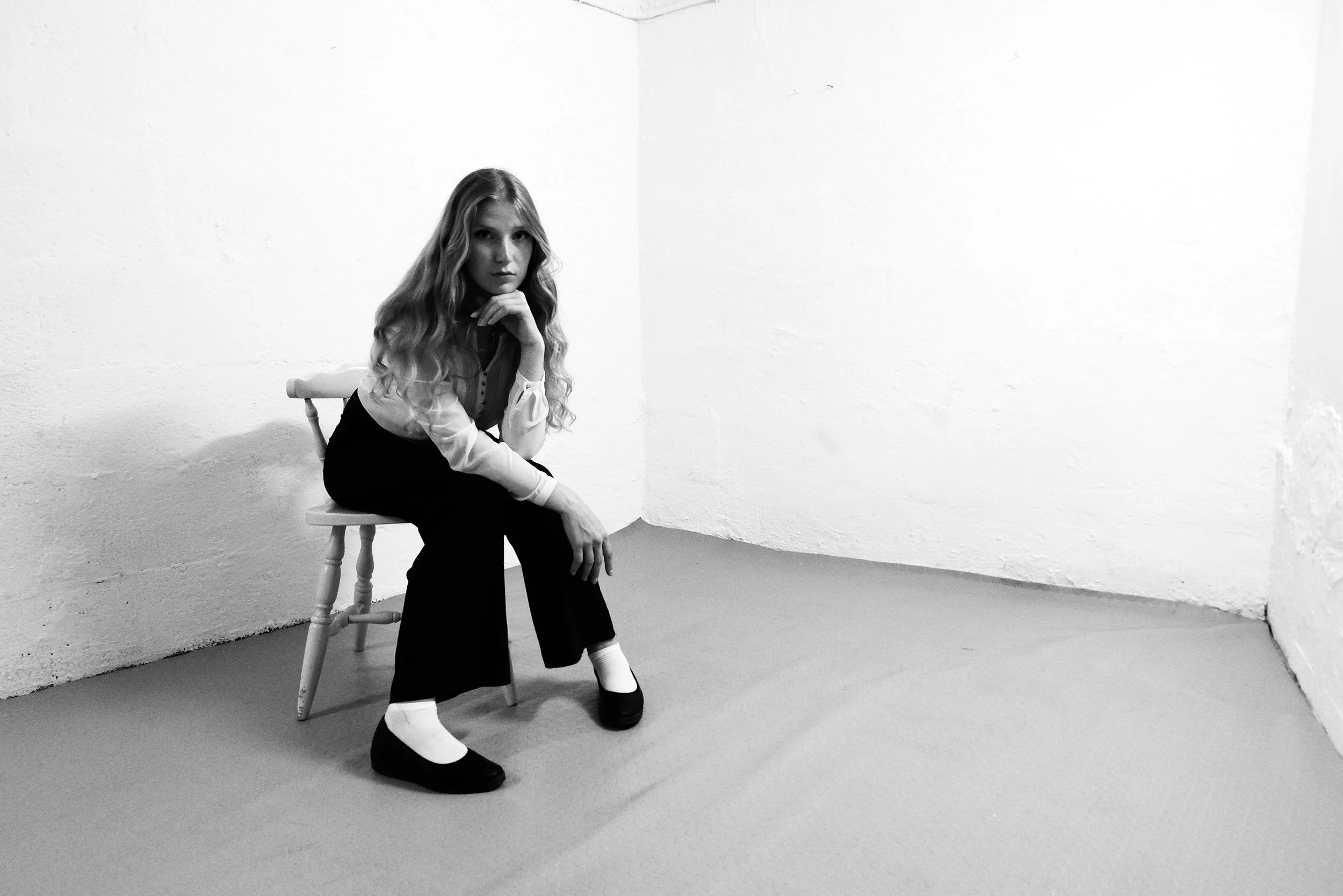 A young woman with long wavy hair sitting on a wooden chair in a corner of a white room, wearing a light-colored blouse, dark pants, and black shoes with white socks, looking at the camera with her chin resting on her hand.