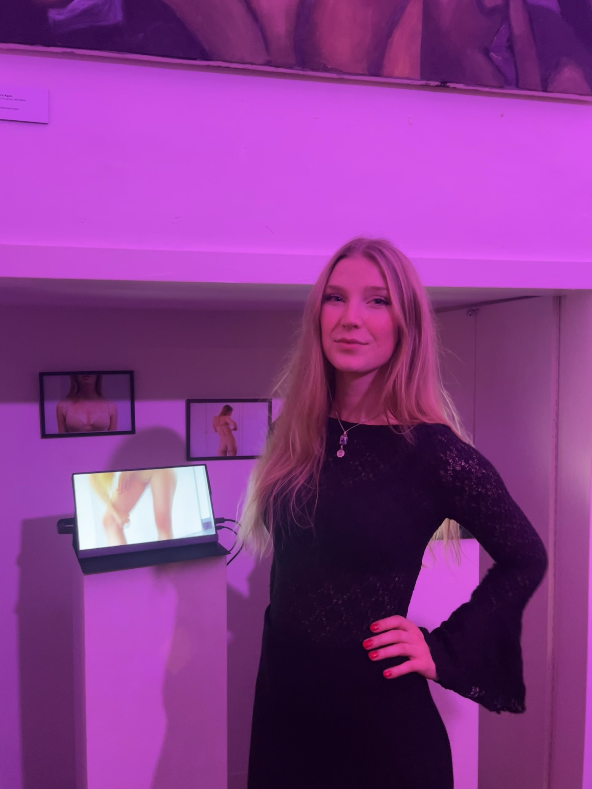 A woman with long blonde hair wearing a black lace dress, standing with one hand on her hip in front of a digital display and framed photographs on the wall, under pink-purple lighting.