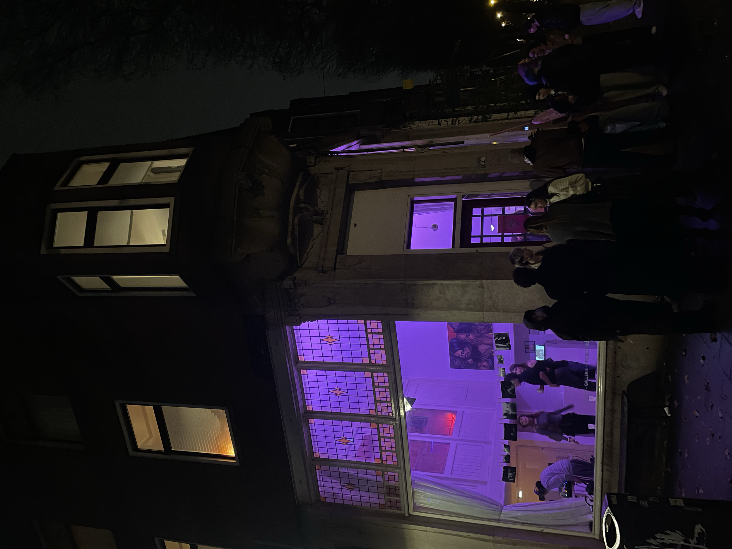 Nighttime view of an art gallery with purple-lit stained glass and people socializing inside, visible through large windows.