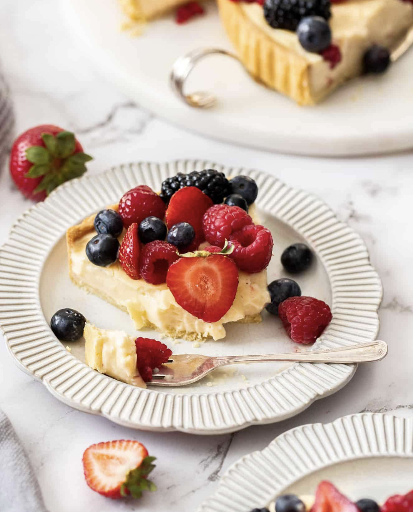 Slice of custard tart topped with strawberries, blueberries, raspberries, and blackberries on a white textured plate.
