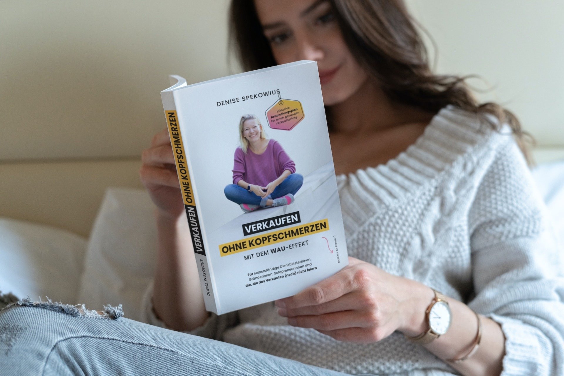 Eine Frau liest ein Buch mit dem Titel "Verkaufen ohne Kopfschmerzen". Das Buch zeigt ein lachendes Mädchen in einem lila Pullover auf dem Cover, sitzt im Schneidersitz.