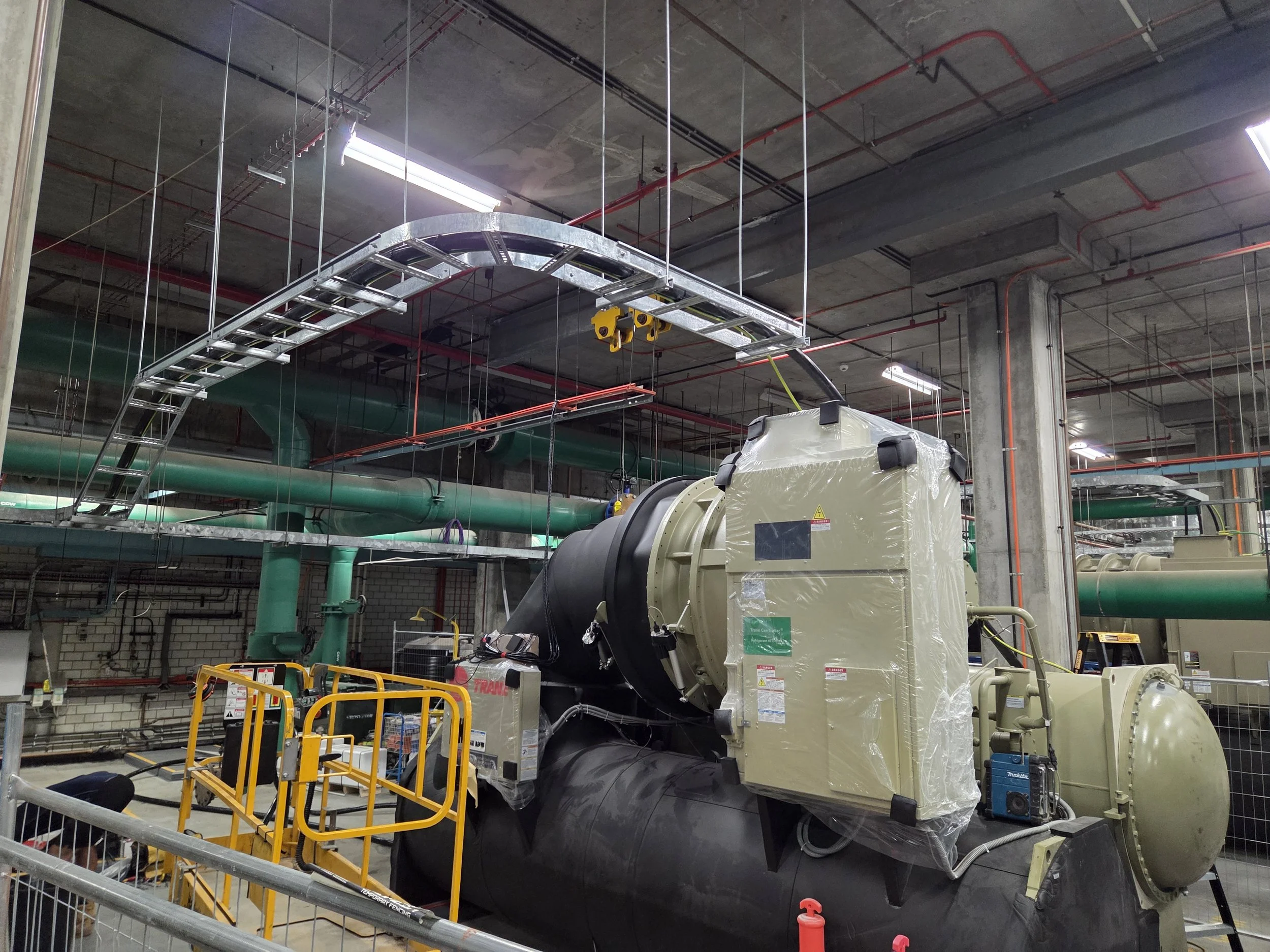 Industrial machinery and equipment in a utility room, including a large generator or engine, surrounded by pipes and metal scaffolding.