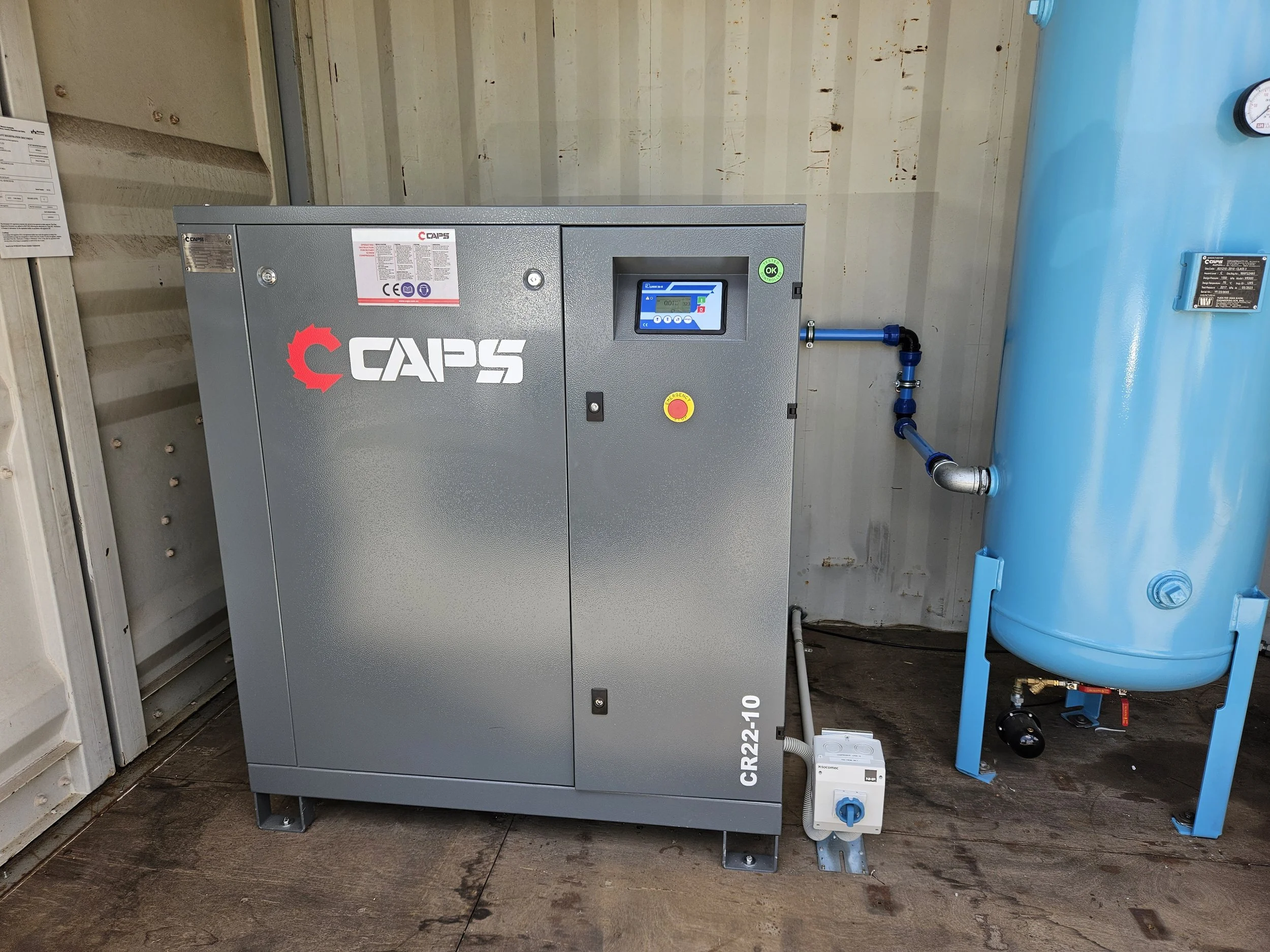 Industrial air compressor system with gray metal cabinet branded 'CAPS' and a large blue tank connected with pipes.