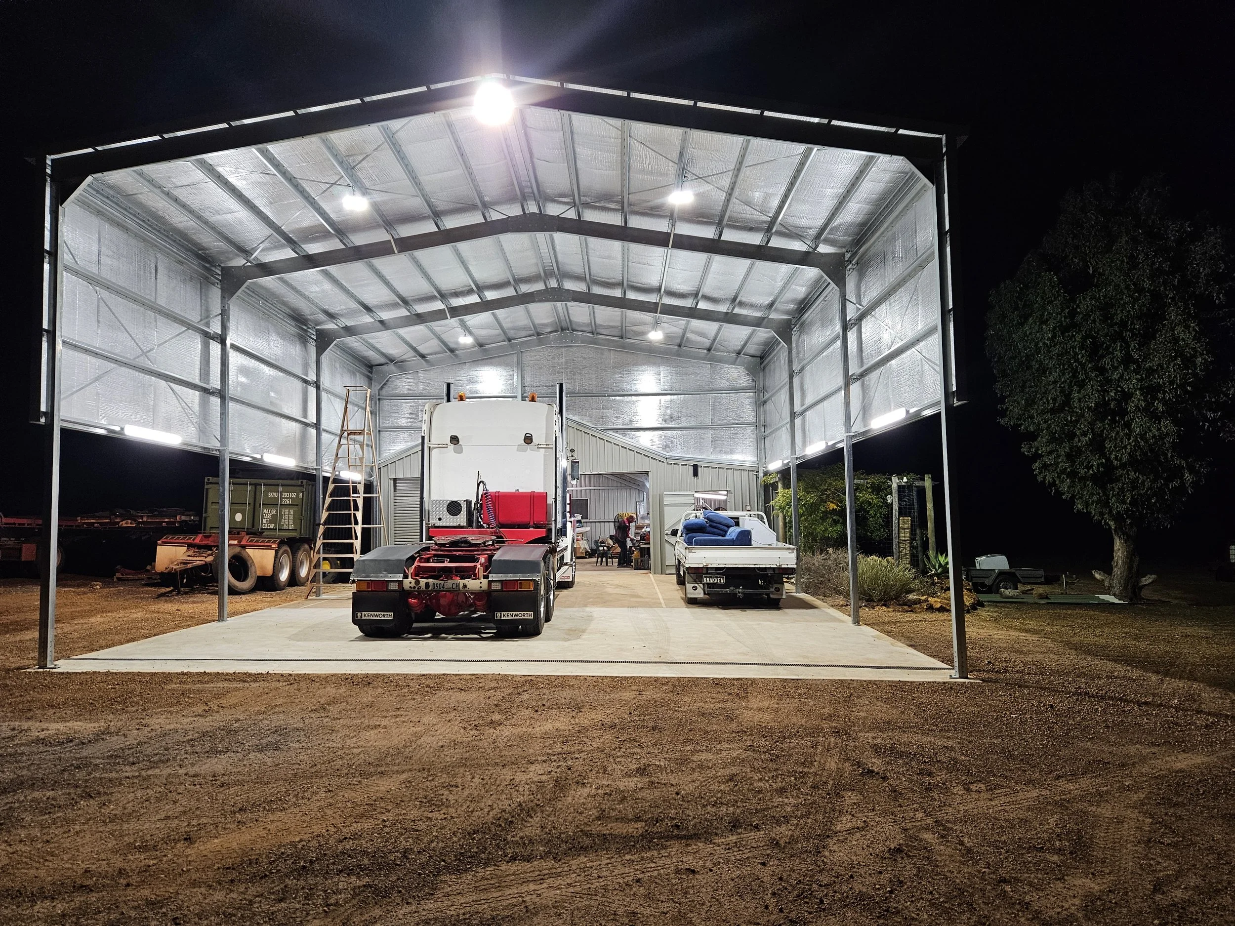 An illuminated metal shed with a truck and a trailer inside at night, surrounded by dirt ground and trees.