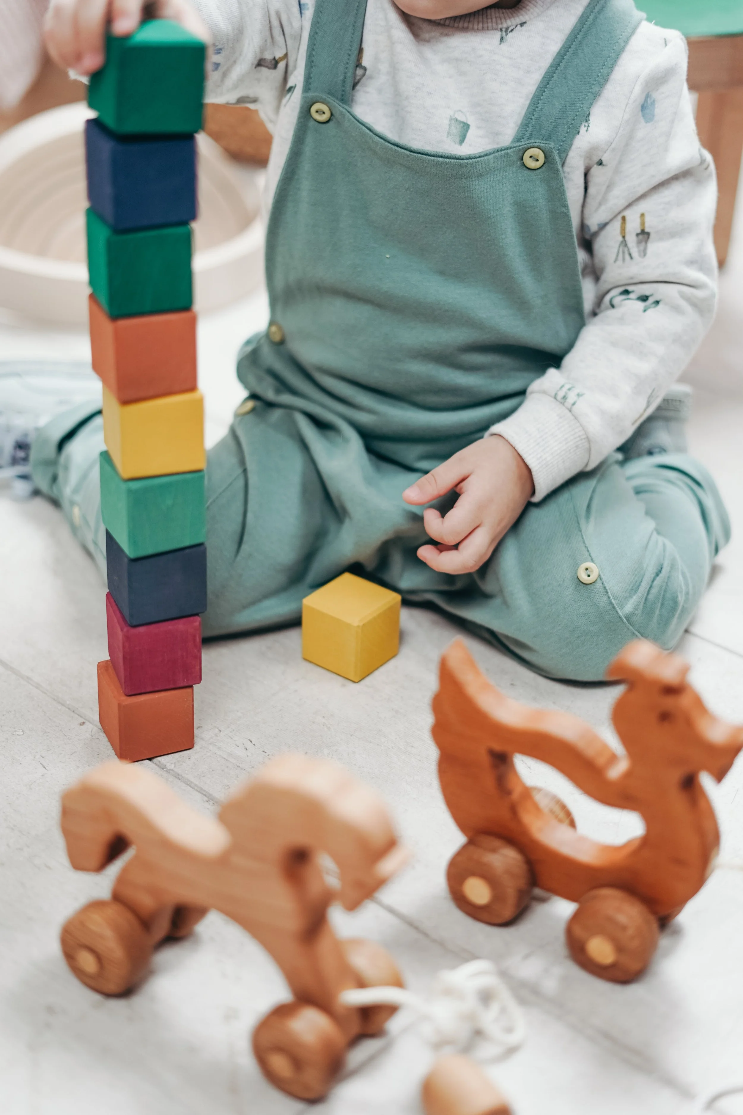 Child sitting on the floor playing with colorful wooden blocks stacked vertically. There are also two wooden toy animals with wheels in the foreground.