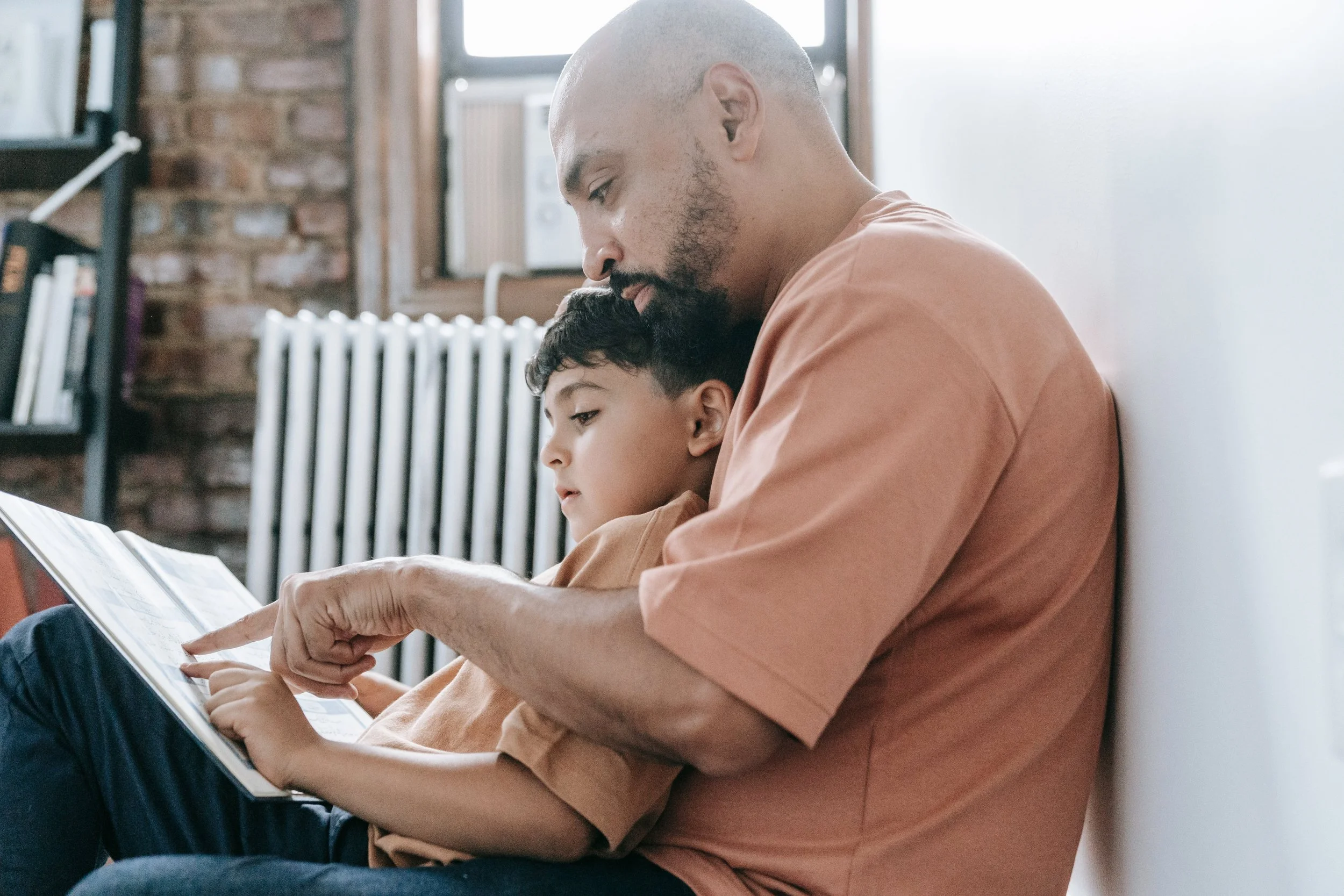 A man and a young boy sitting on a bench, looking at a book together, in a room with brick walls and a heater.
