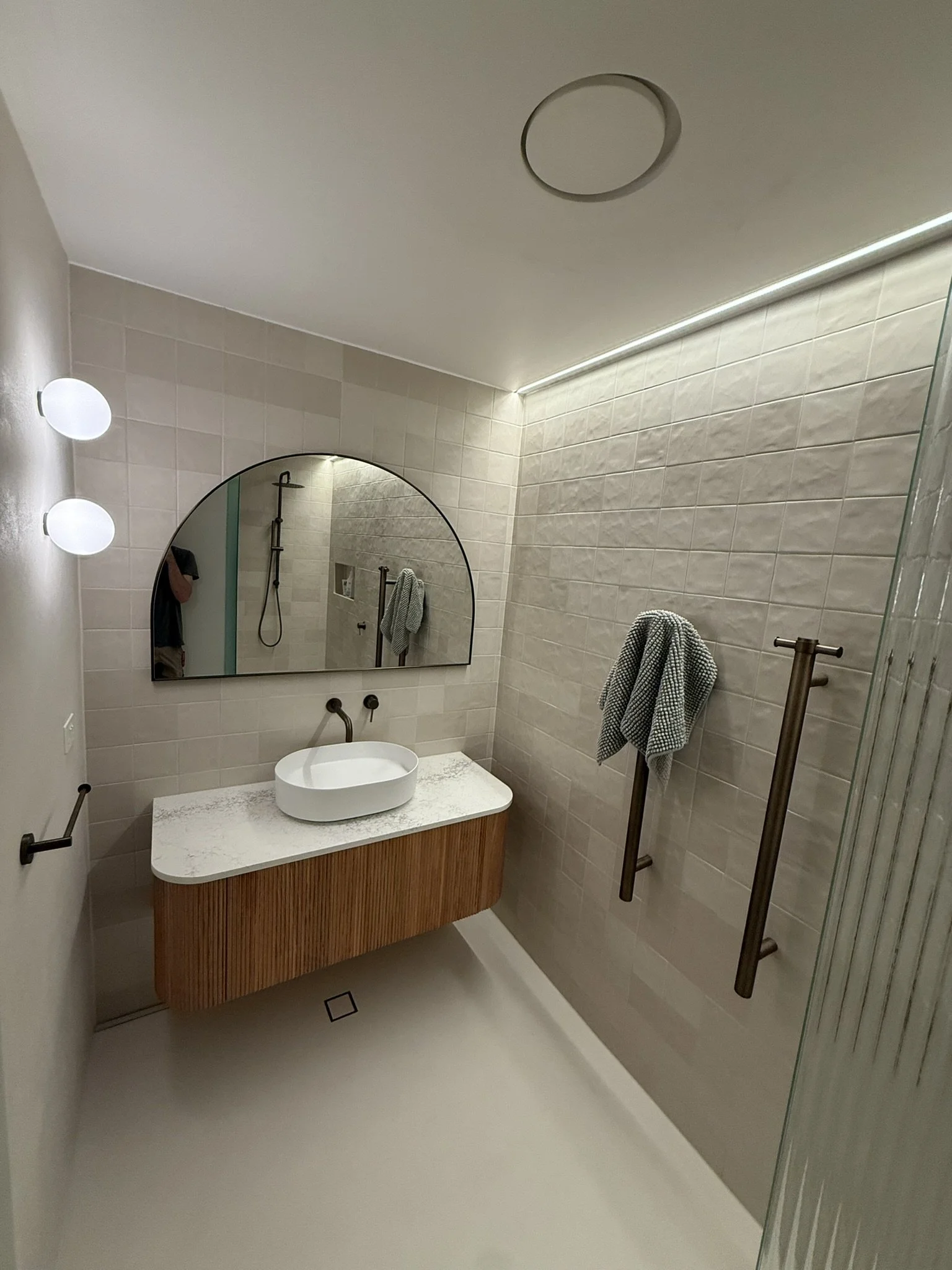 Modern bathroom with beige tiled walls, a large round mirror, a vessel sink on a wooden cabinet, a towel hanging on a black towel rack, wall-mounted lights, and a walk-in shower with a showerhead and glass partition.