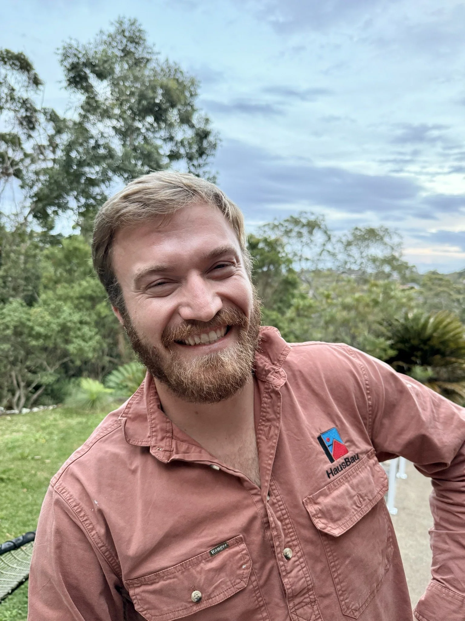 a smiling man with a beard and mustache wearing a salmon-colored shirt with a logo on the pocket, standing outdoors with trees and a cloudy sky in the background