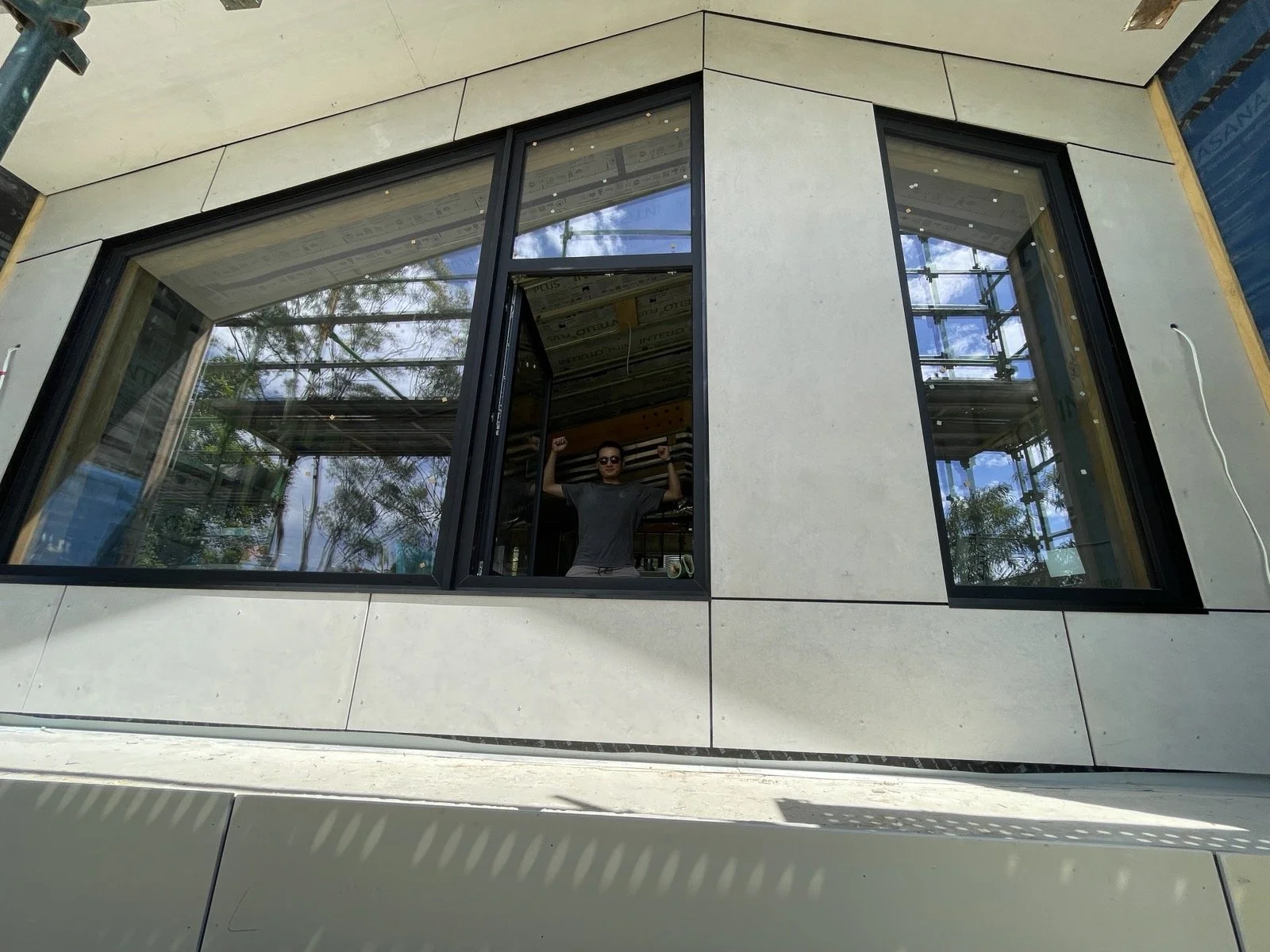 A construction worker standing inside a large window opening of a building under construction, with trees and scaffolding visible outside.