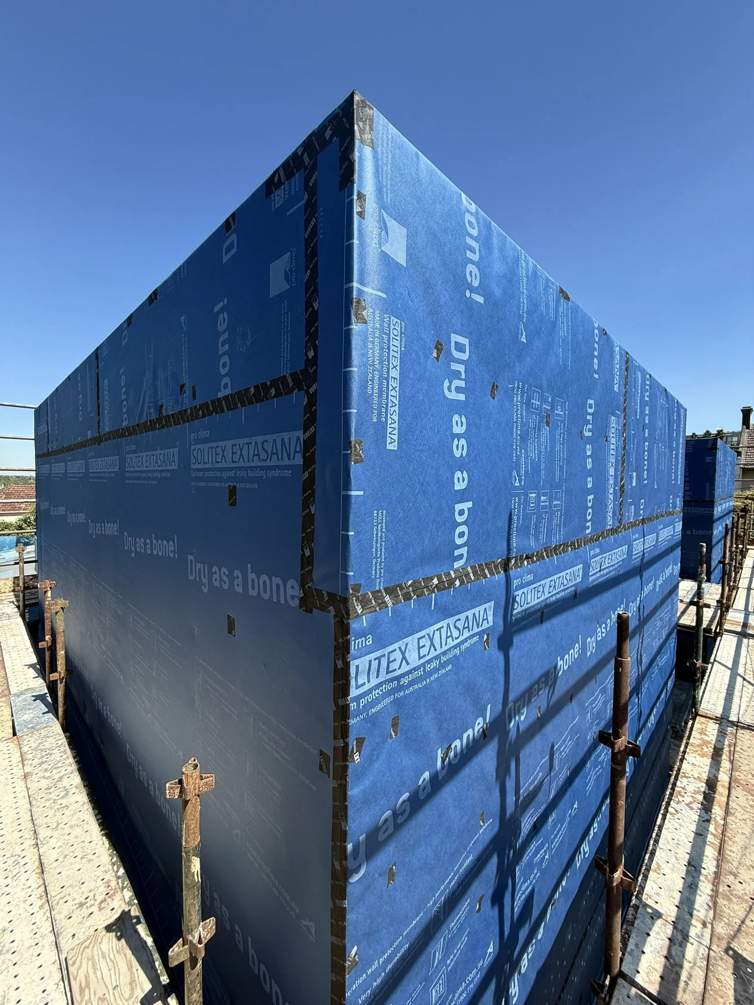 Construction site with a building covered in blue insulation panels and scaffolding, against a clear blue sky.