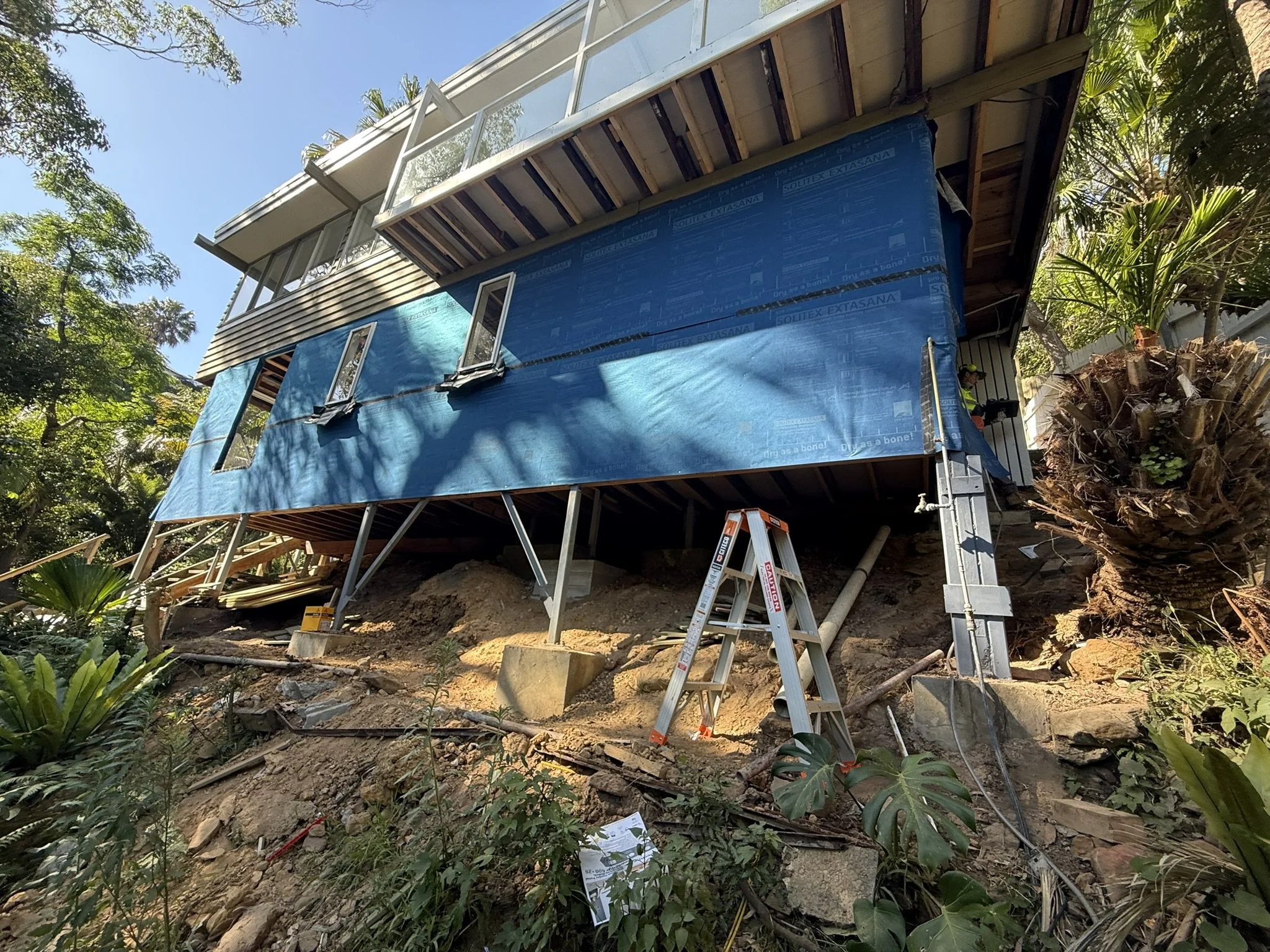 A house under construction on a hillside with scaffolding and blue weather-resistant sheeting, surrounded by trees and plants.