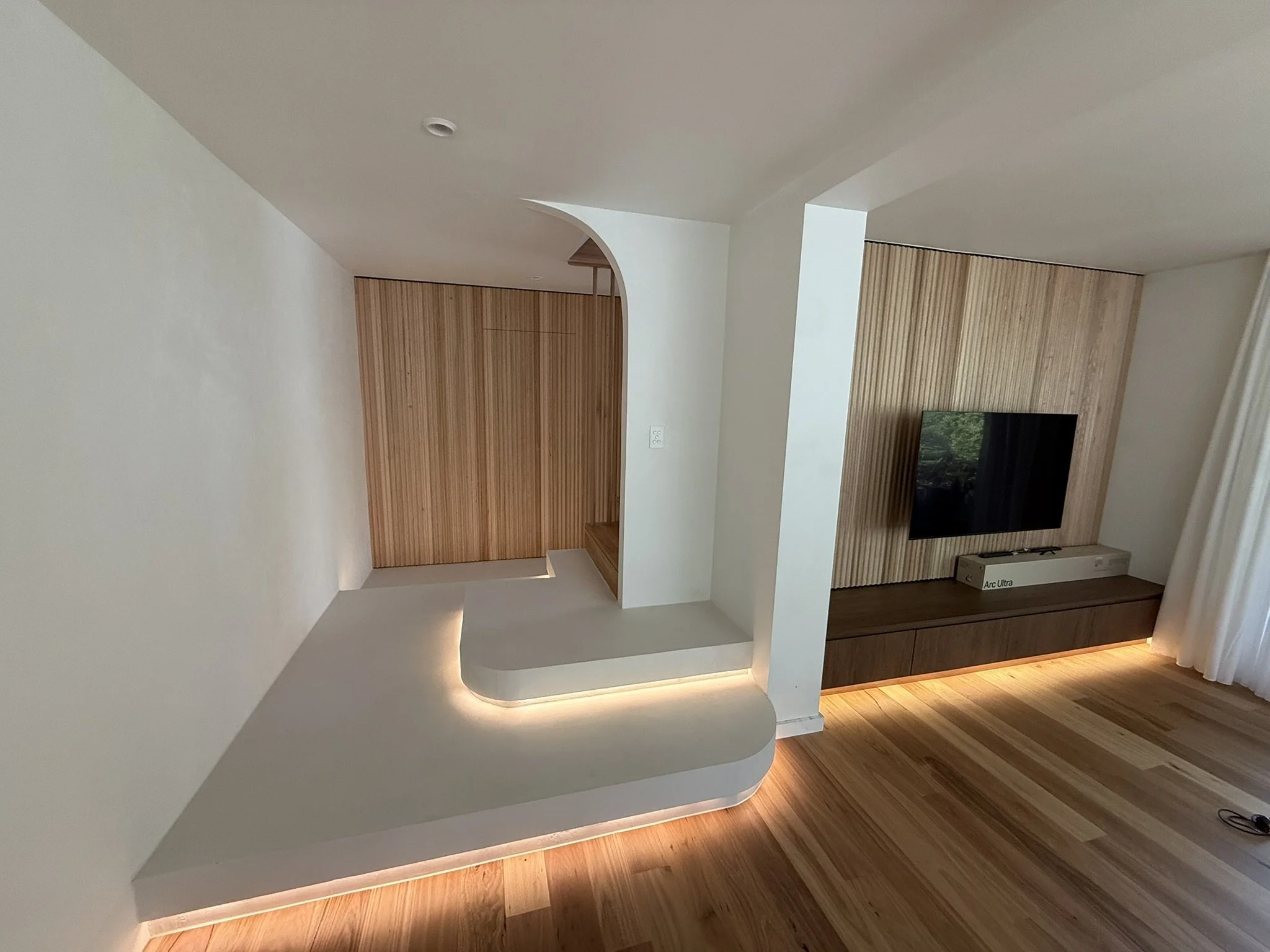 Modern living room with wooden panel accent wall, mounted flat-screen TV, white curved architectural feature, and ambient lighting along the base of the wall.