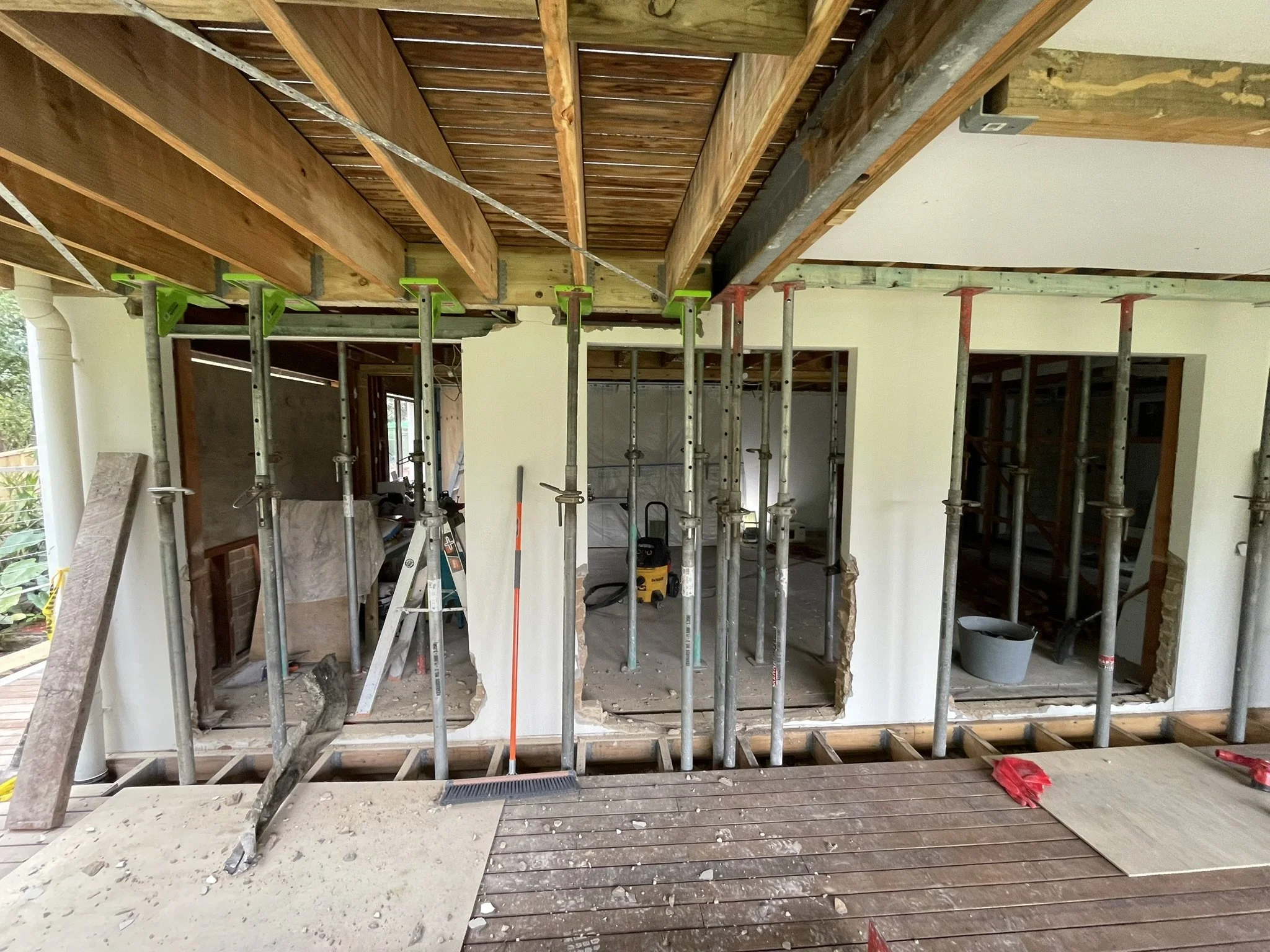 The image shows a room under construction with scaffolding in the center, a partially removed wall, exposed wooden beams and ceiling, construction tools, and debris on the floor.
