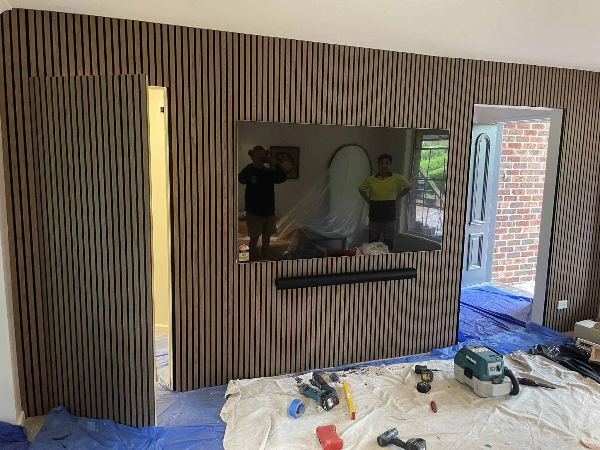 Interior view of a room under renovation with a wall covered in vertically arranged wood panels, a mounted flat-screen TV, large windows, and construction tools and materials scattered on the floor covered with protective sheets.
