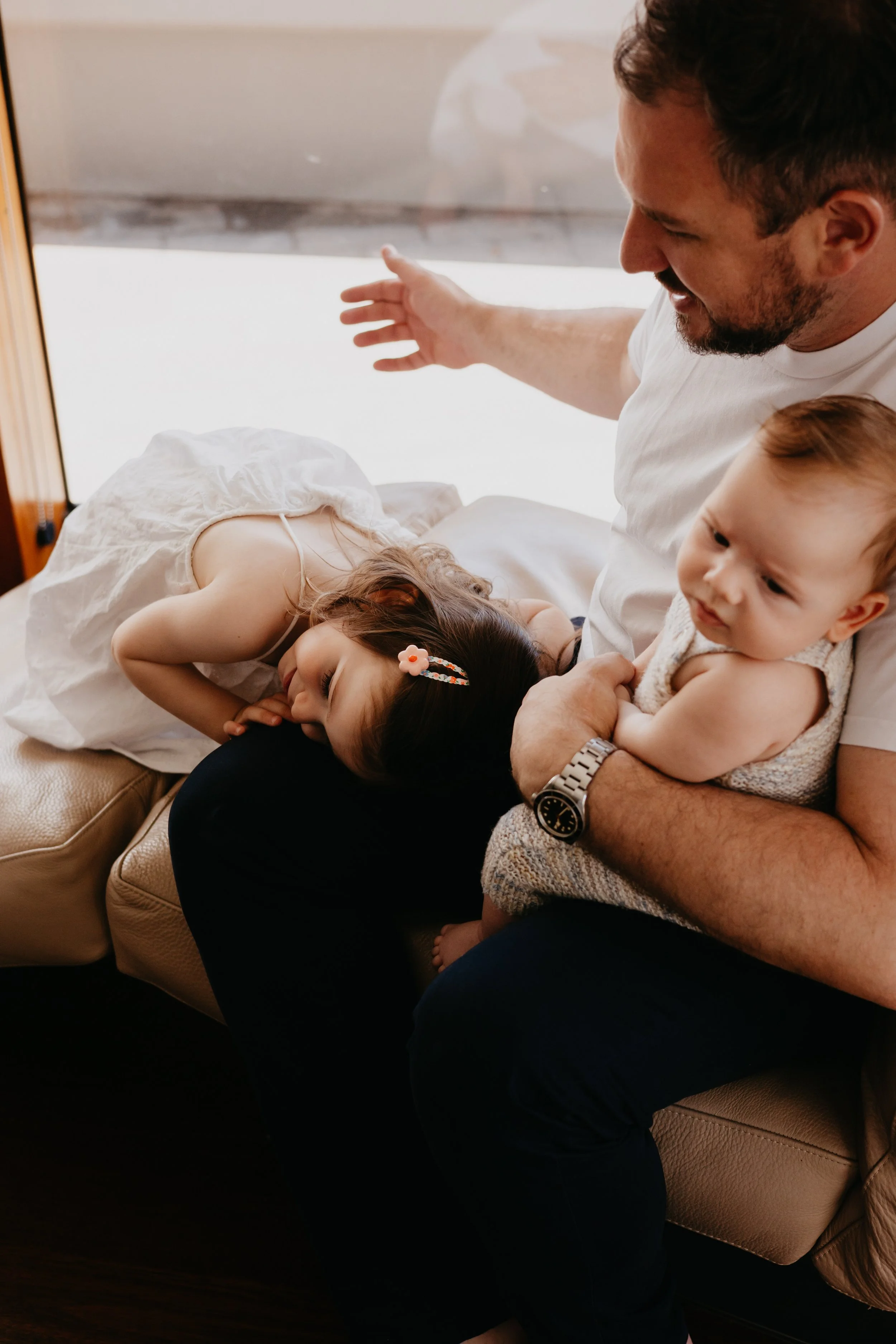 A man sitting on a couch holding a baby in his lap while a young girl, lying on her side on the couch, sleepily rests her head on his knee. The girl is wearing a white dress and a hair clip, and the baby is dressed in a knitted outfit.