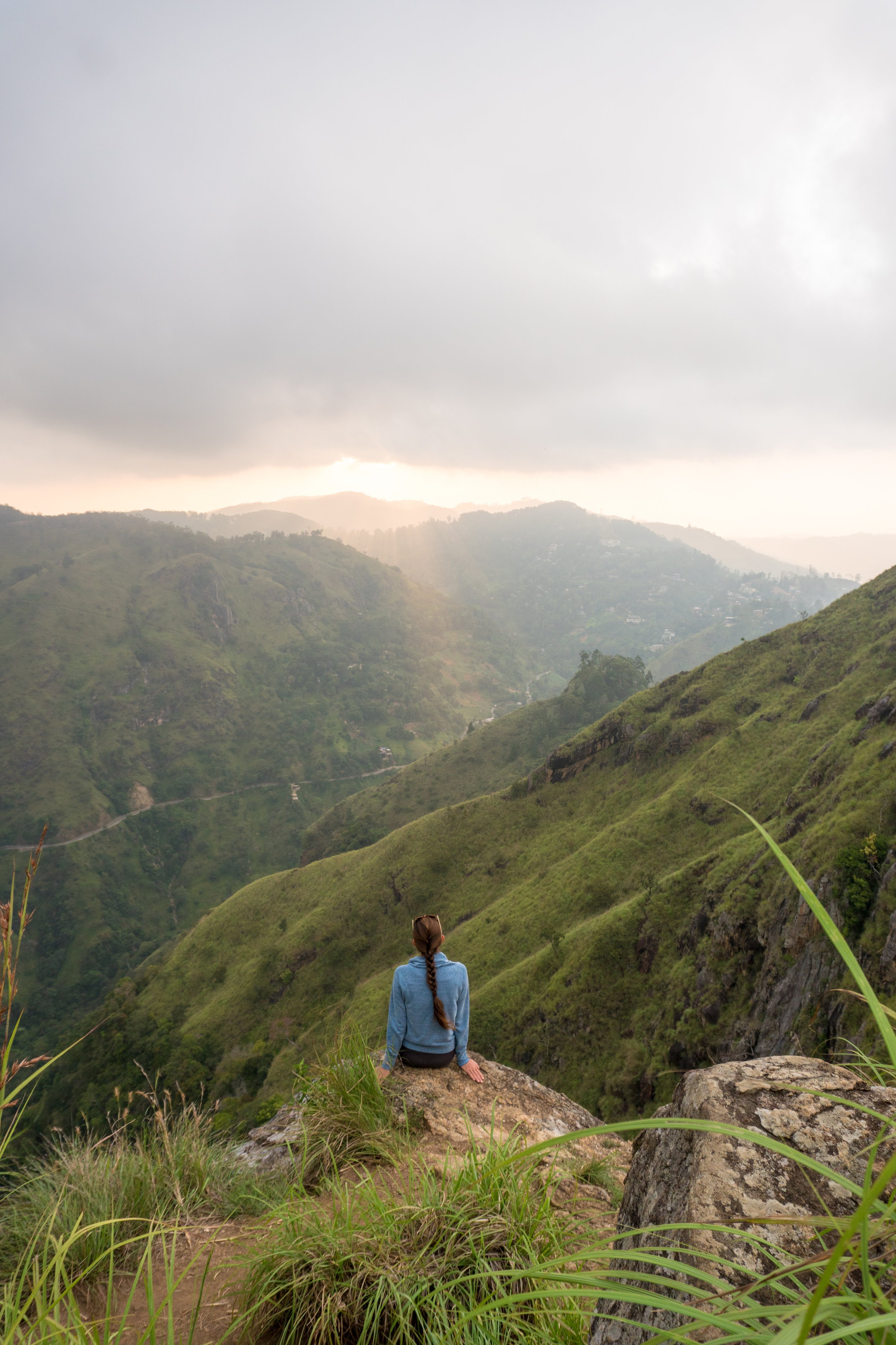 A person sitting on a large rock overlooking lush green mountains and valleys during a cloudy day with rays of sunlight breaking through the cloud cover.