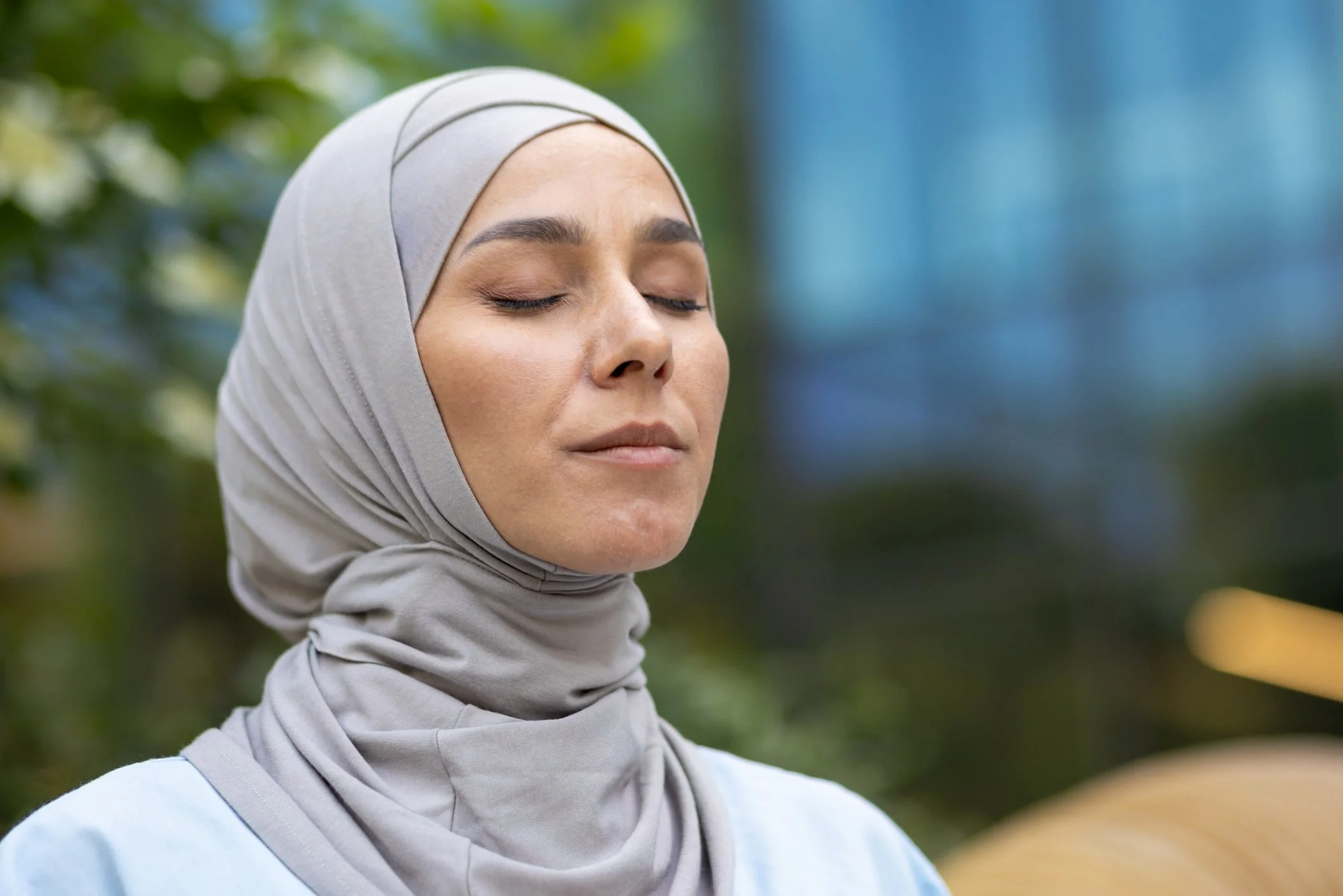 A woman wearing a light grey hijab with closed eyes, standing outdoors with blurred greenery and a building in the background.